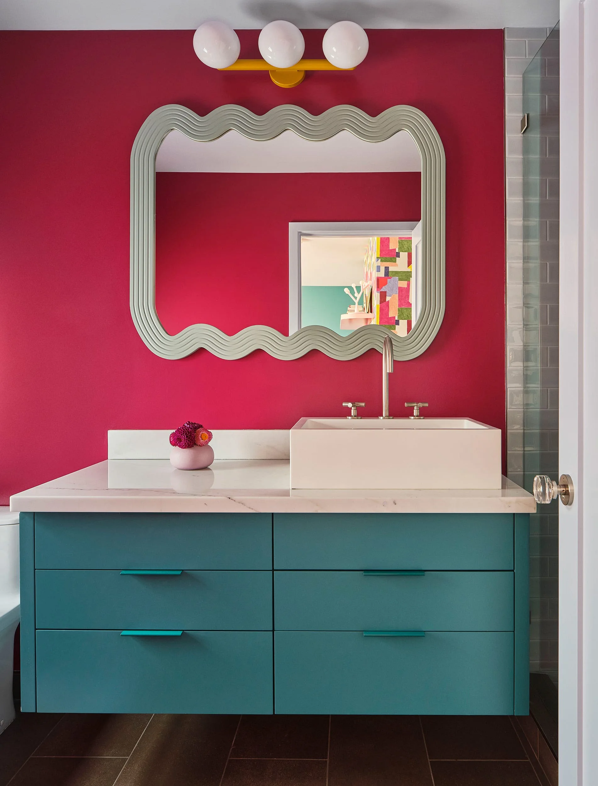 A modern teal floating vanity with a marble countertop, a white vessel sink, a silver wavy mirror, and a magenta painted wall.
