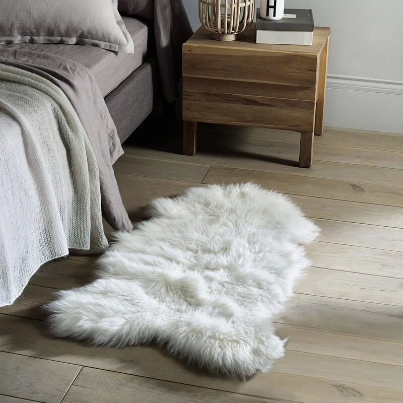 A white faux fur rug lies on light wood flooring next to a wooden nightstand and a bed with grey linen sheets.