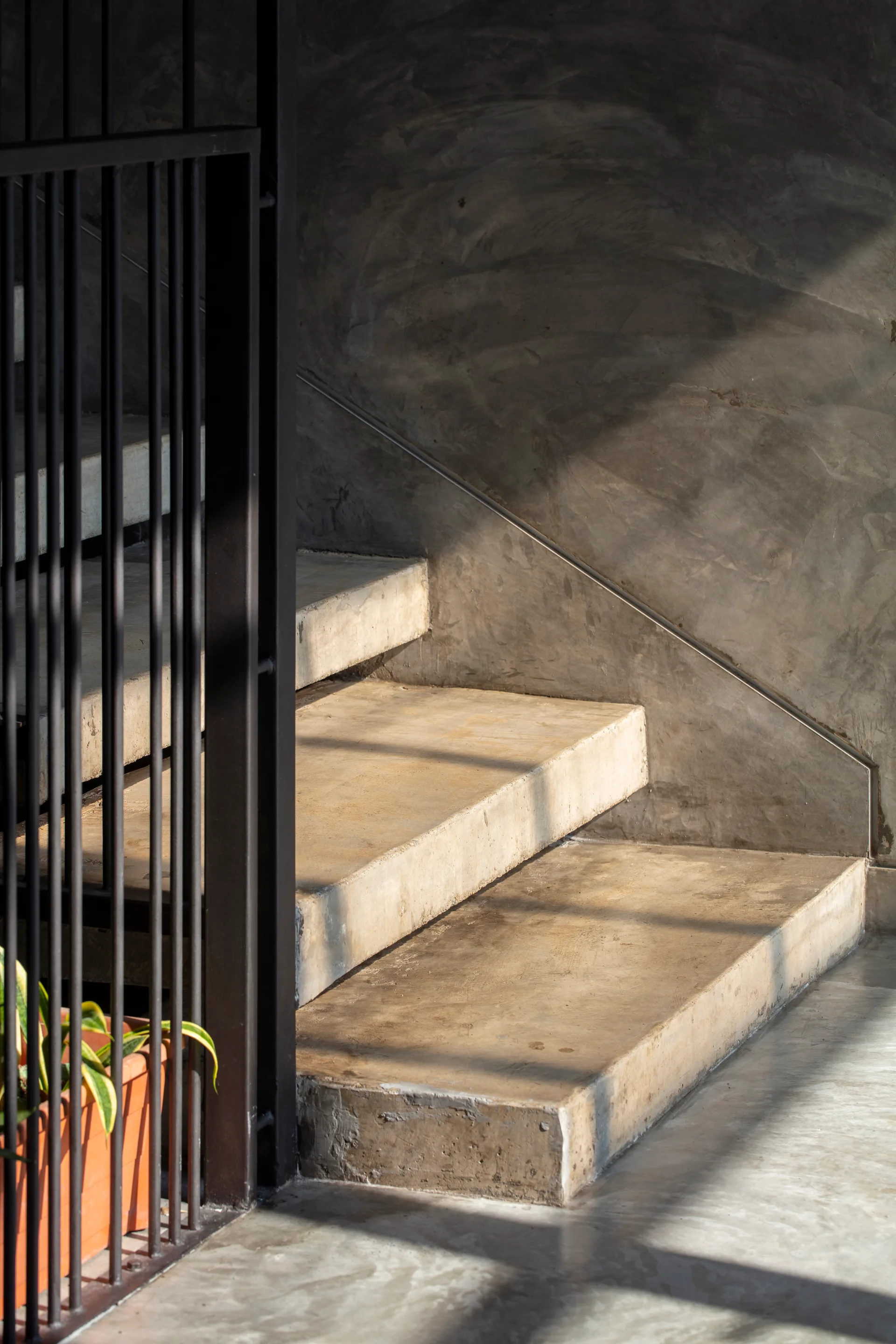 A close-up of minimalist cast concrete steps accompanied by a black vertical metal railing under directional sunlight.