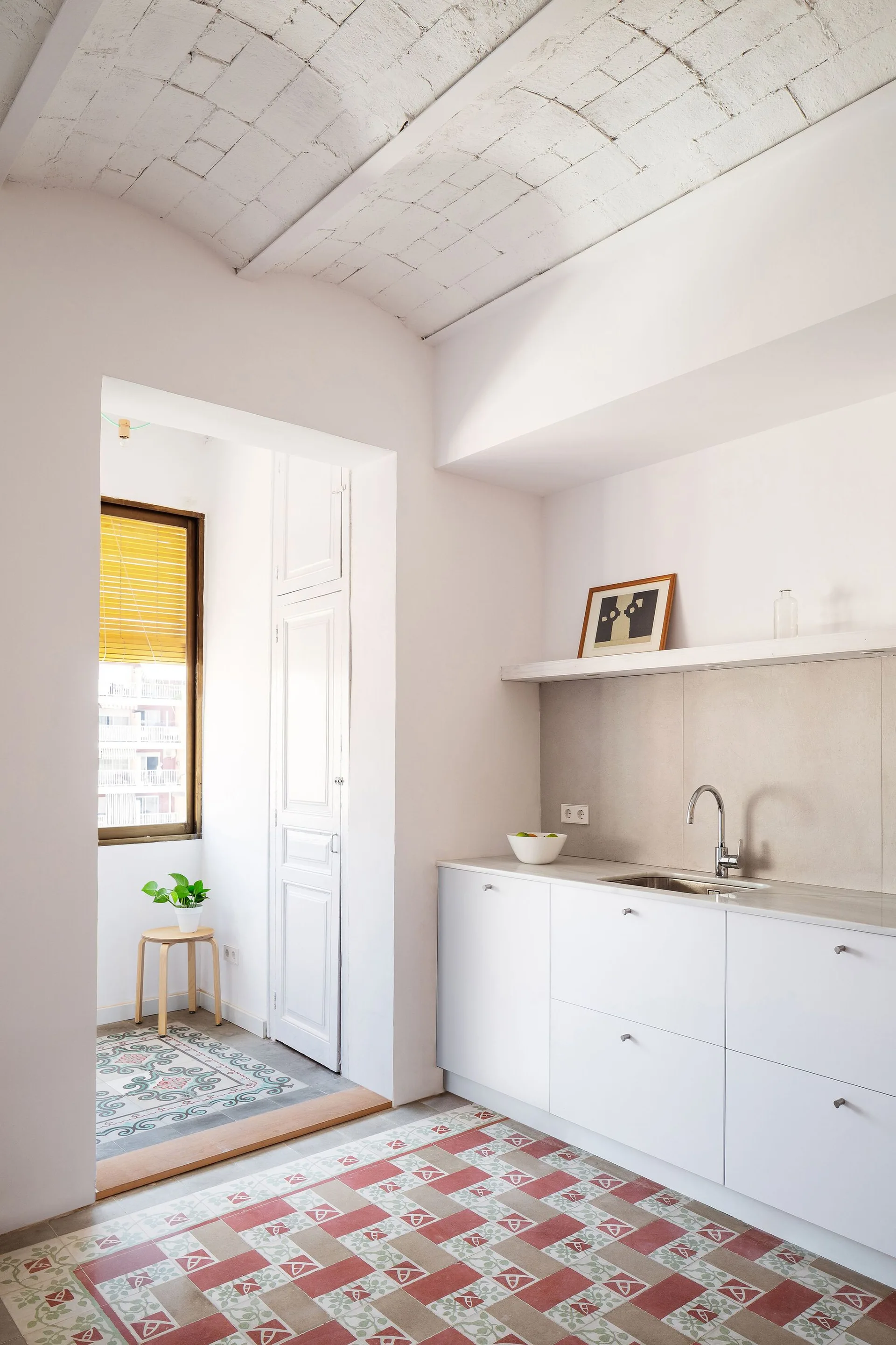 A bright white kitchen features minimal base cabinets, a stainless steel sink, patterned tile flooring, and a whitewashed brick vaulted ceiling.