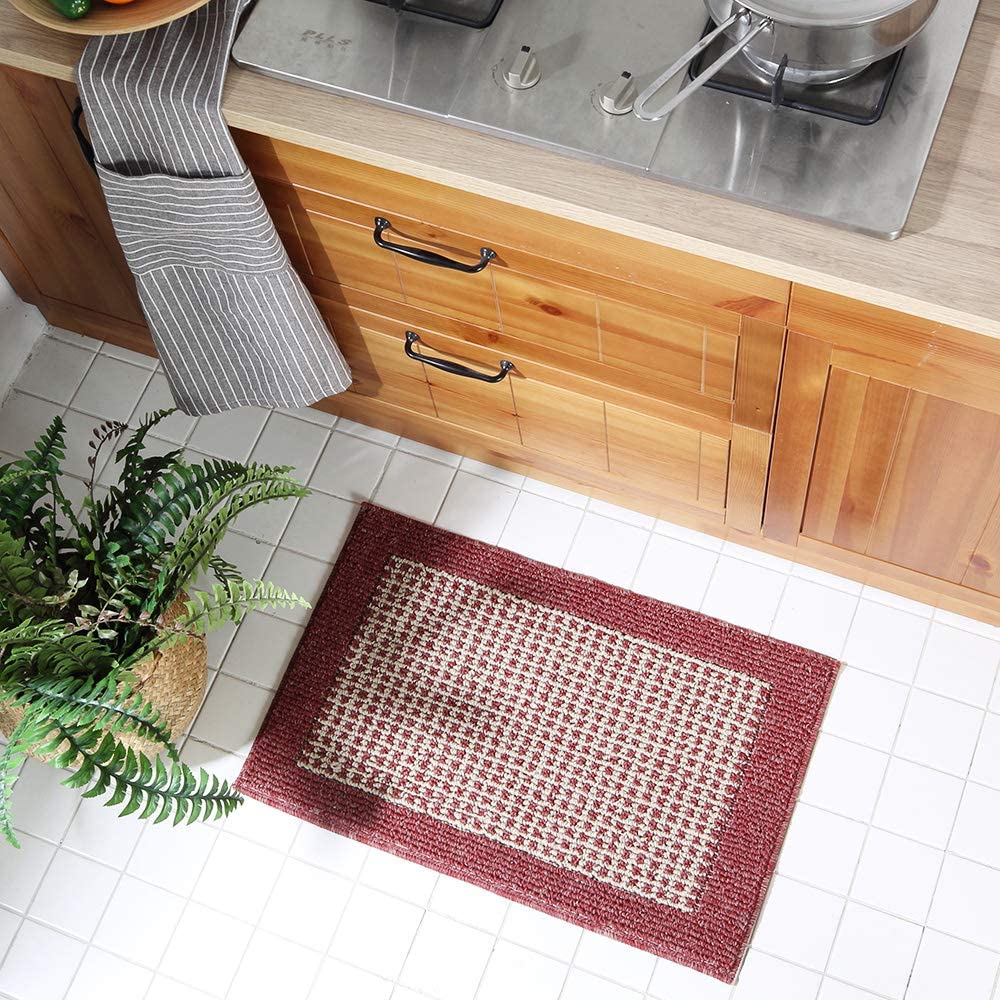 A red and cream checkered woven rug is placed on a white tiled kitchen floor next to wooden cabinets and a green potted plant.