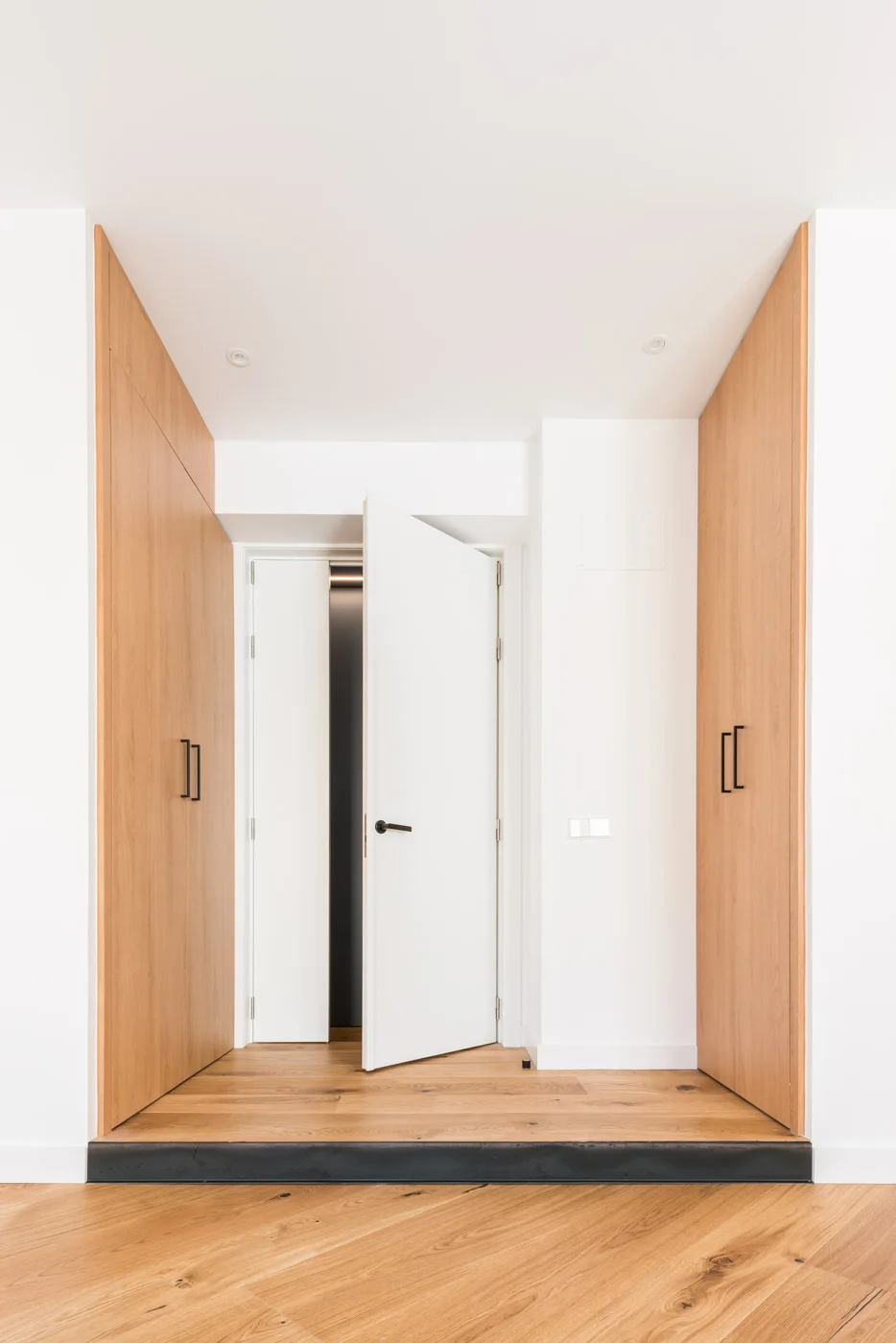 A minimalist entry hall features light oak flooring, floor-to-ceiling wood closets with black handles, and a partially open white door.