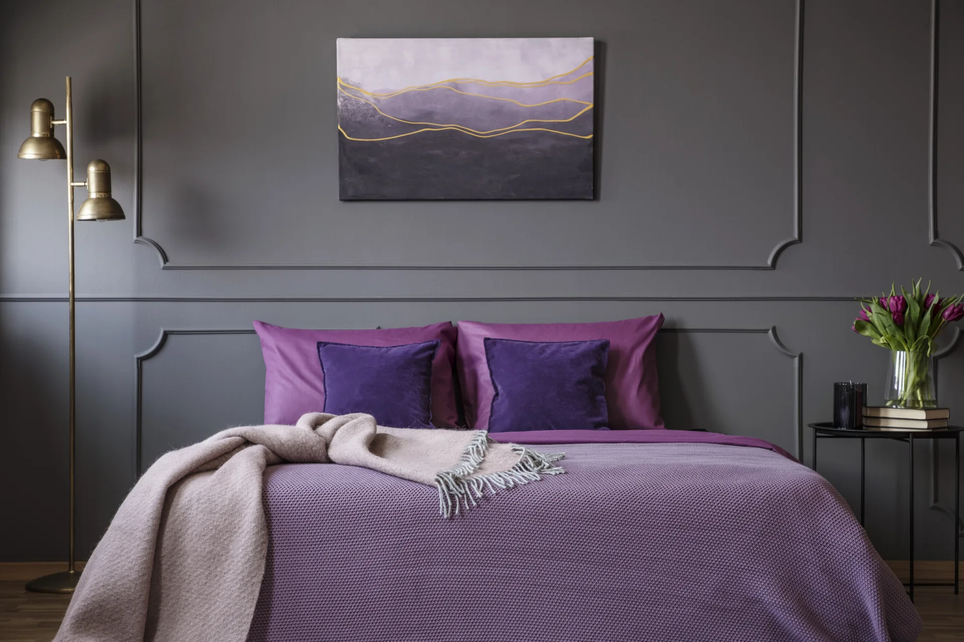 A bedroom features charcoal gray walls with rectangular molding, purple bedding with velvet pillows, a gold floor lamp, and abstract wall art.