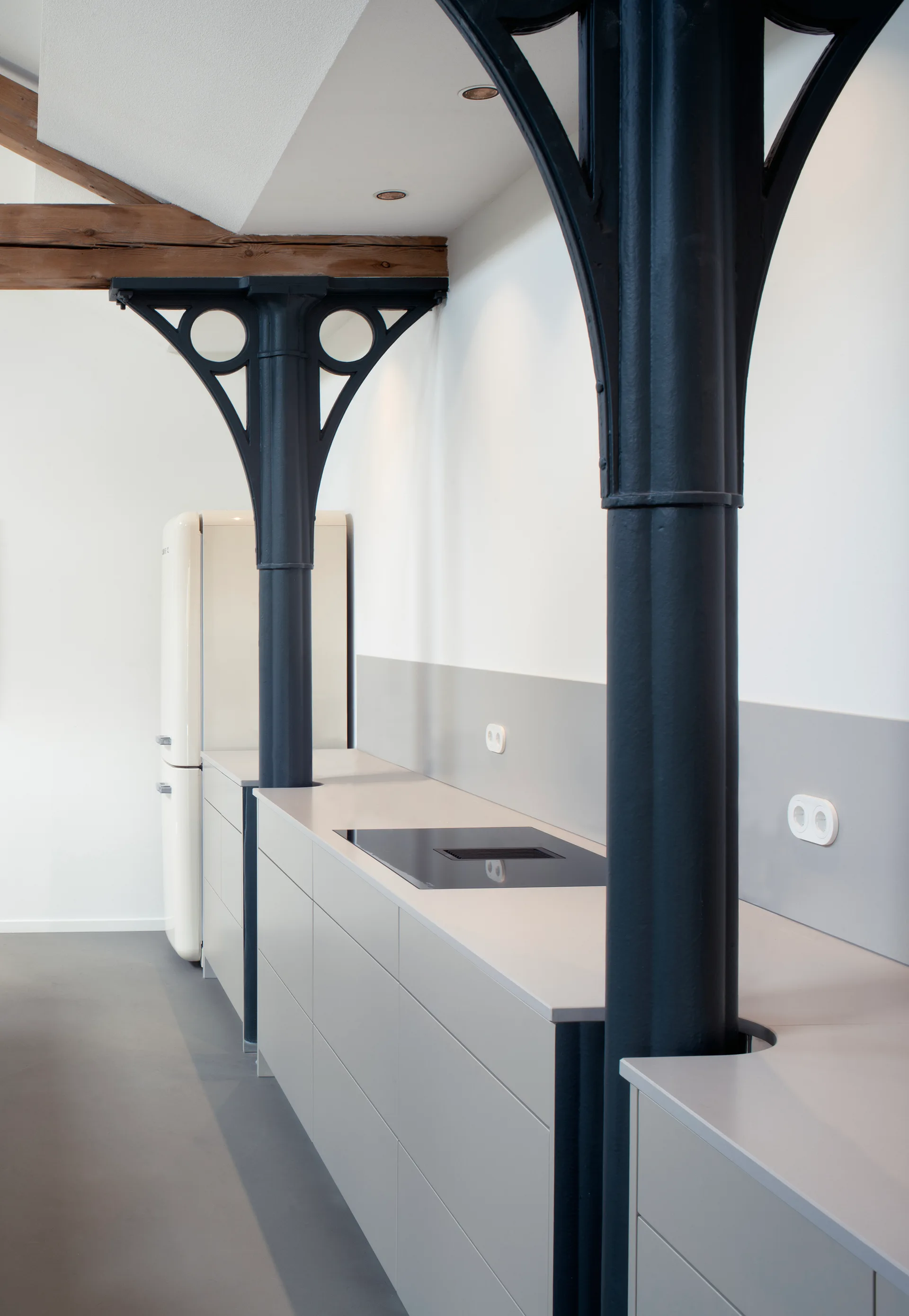 Dark iron columns integrated into a light gray kitchen countertop with minimalist cabinets and a retro cream refrigerator.
