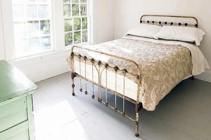 A light-filled bedroom features an antique metal bed frame with patterned bedding next to a white multi-paned window and a painted green dresser.