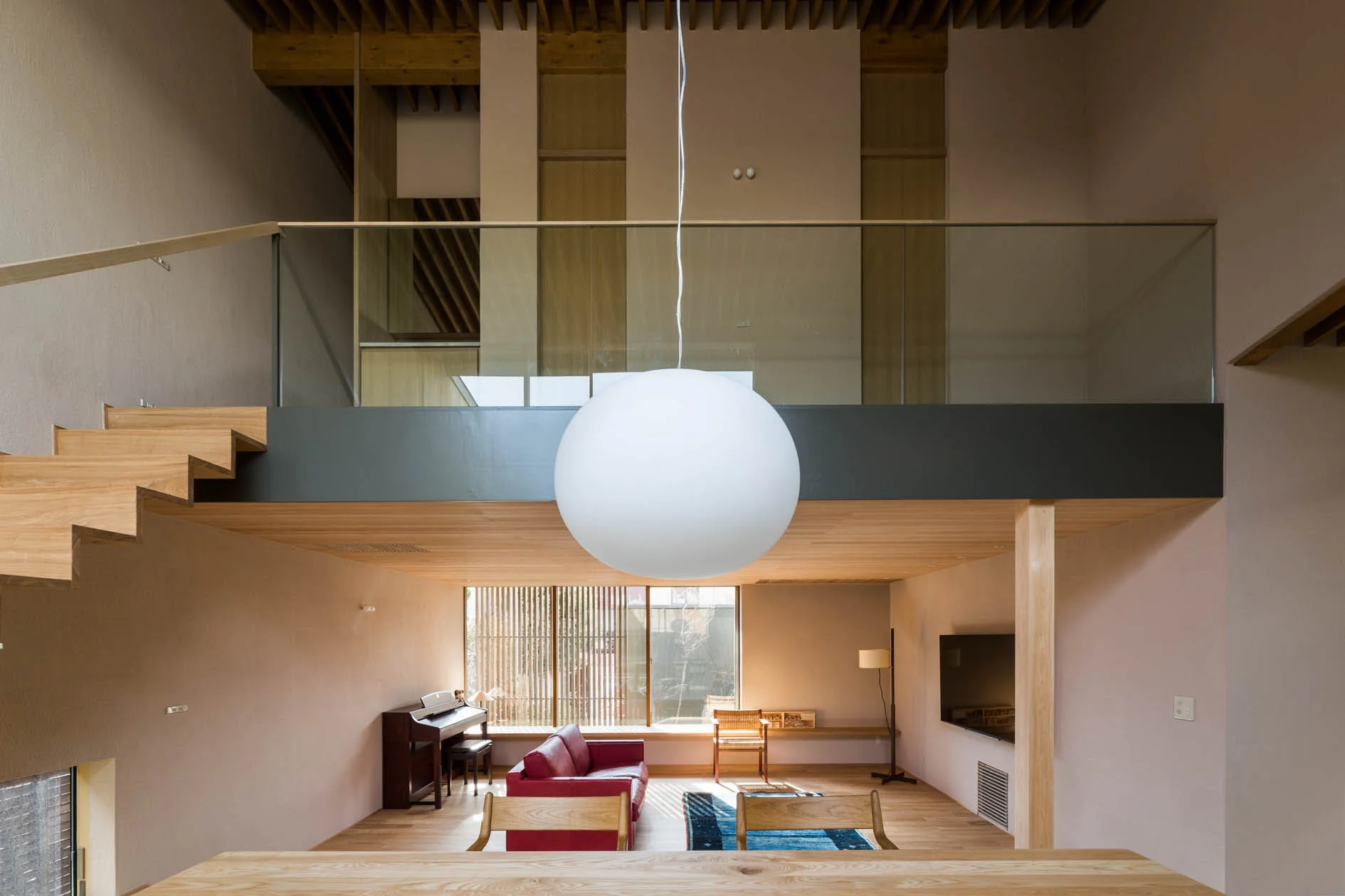 A split-level living room features a red leather sofa, a floating wooden staircase, a glass-railing mezzanine, and a large white globe pendant light.