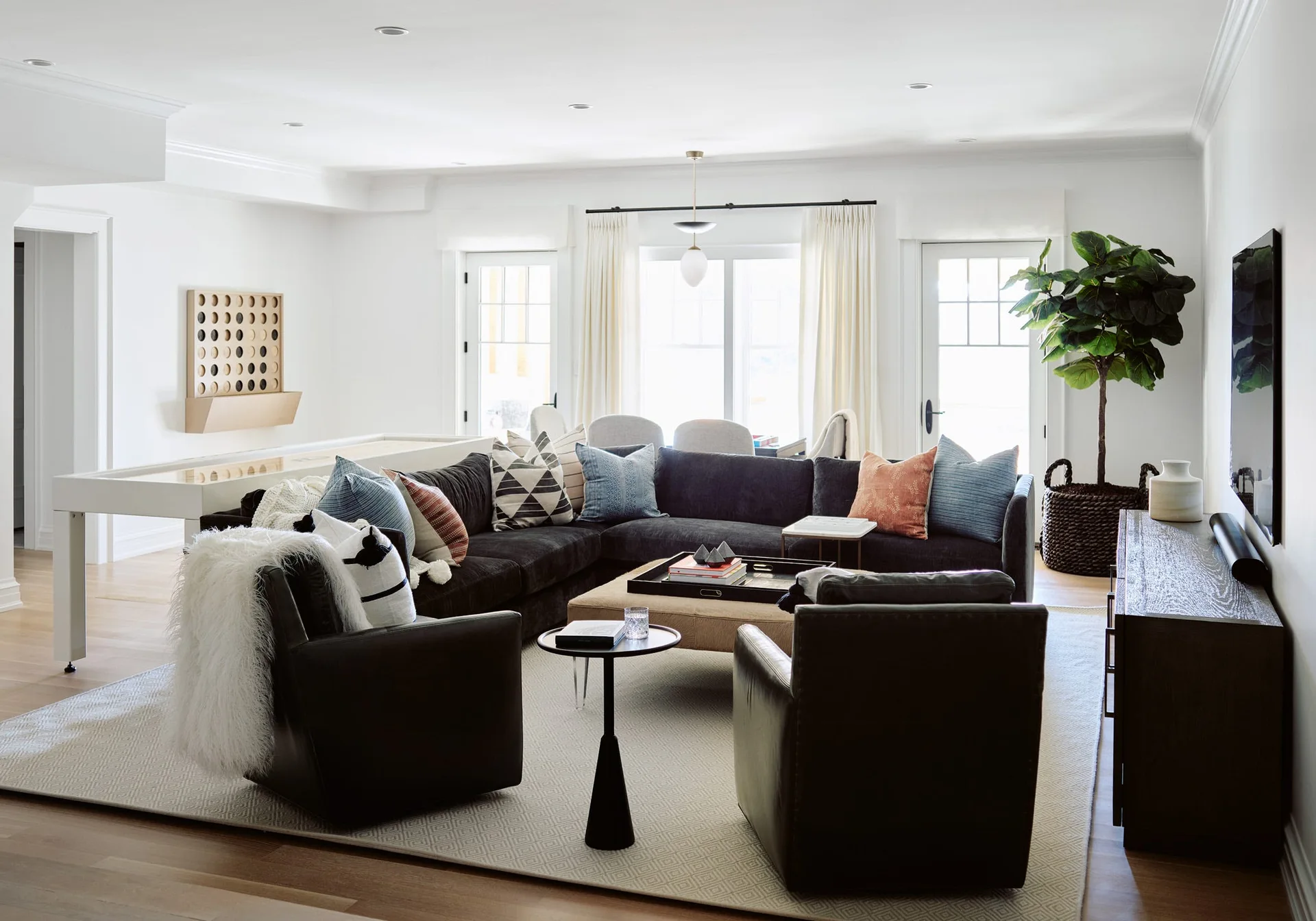 A spacious white living room featuring a dark charcoal sectional, two leather armchairs, an upholstered coffee table, and a white shuffleboard table.