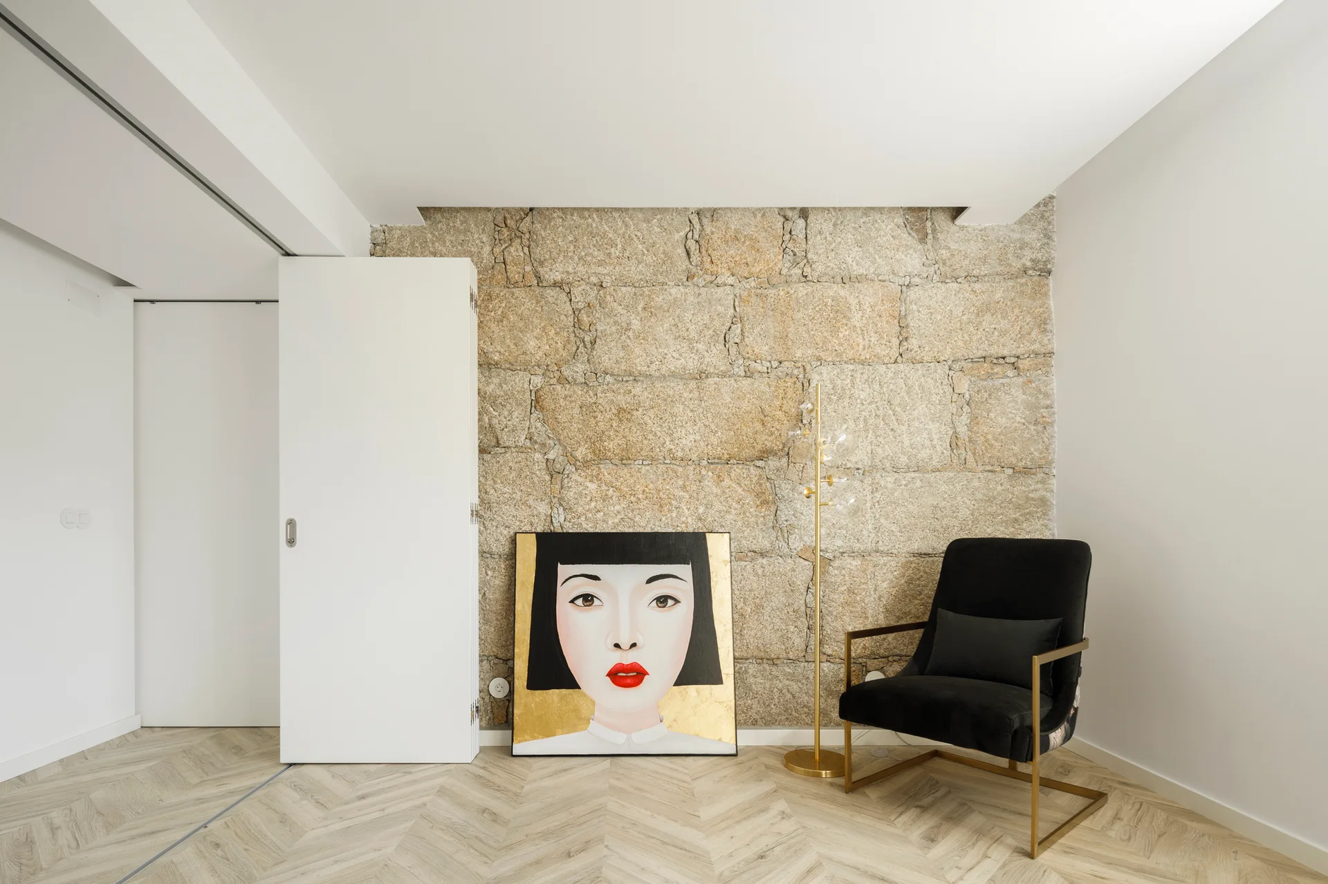 A minimalist room corner features a textured stone wall, chevron patterned wood floors, a black armchair, and a large portrait painting on the floor.