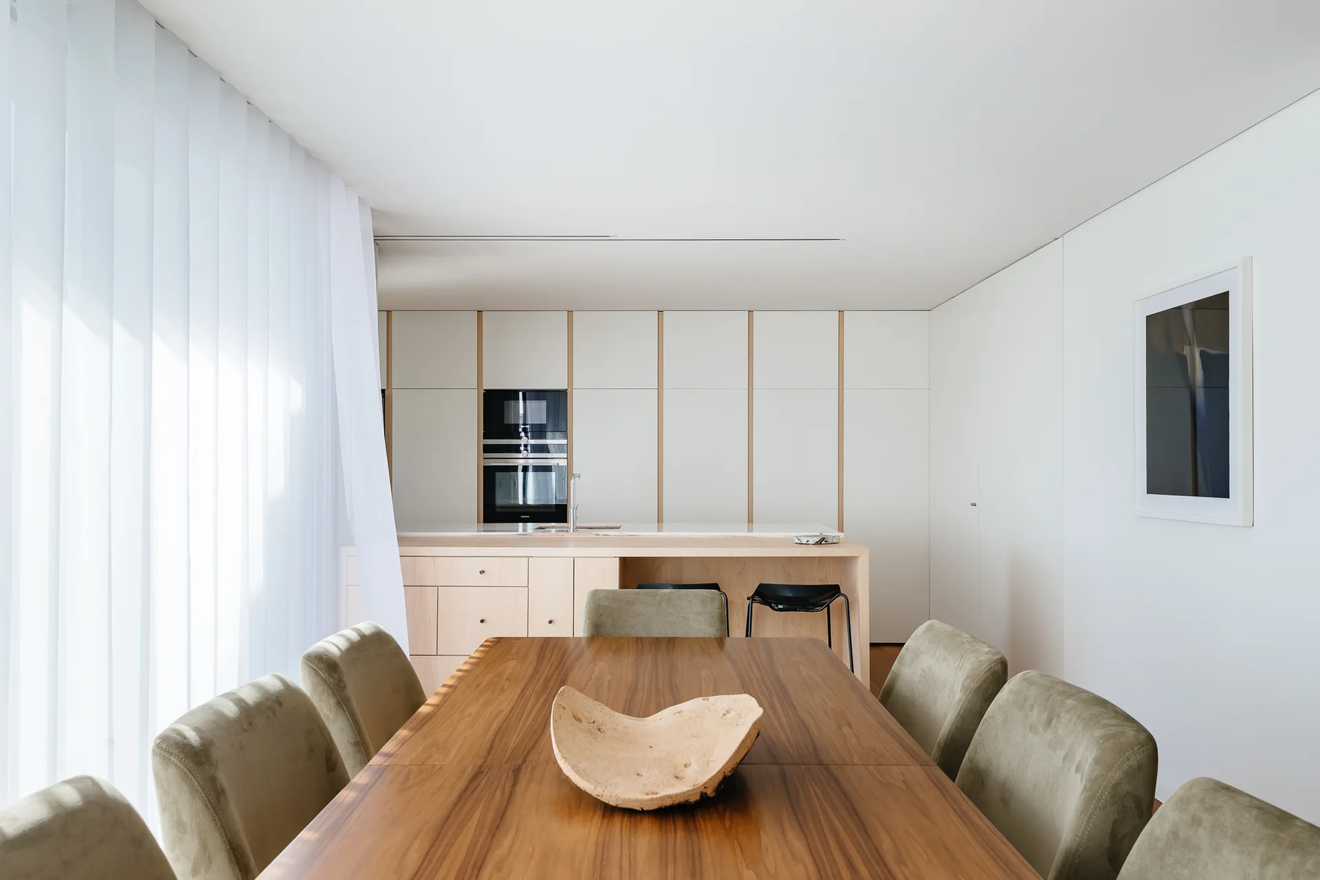 A modern dining room features a warm wood table with olive green upholstered chairs, opening to a minimalist white kitchen with sheer curtains.