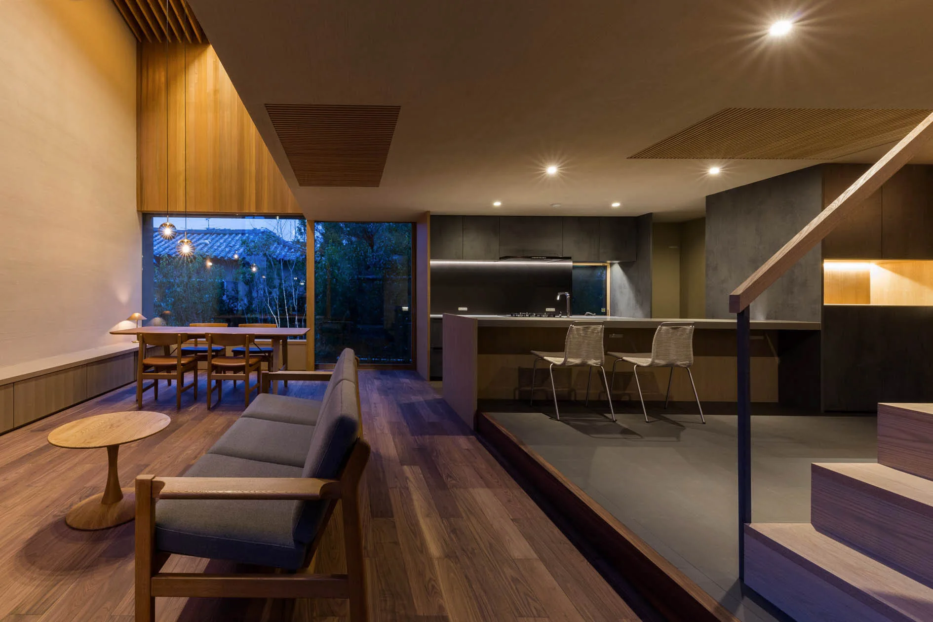 A modern open-concept home features a wood-floored living room with a gray sofa and a raised platform kitchen with dark cabinetry.