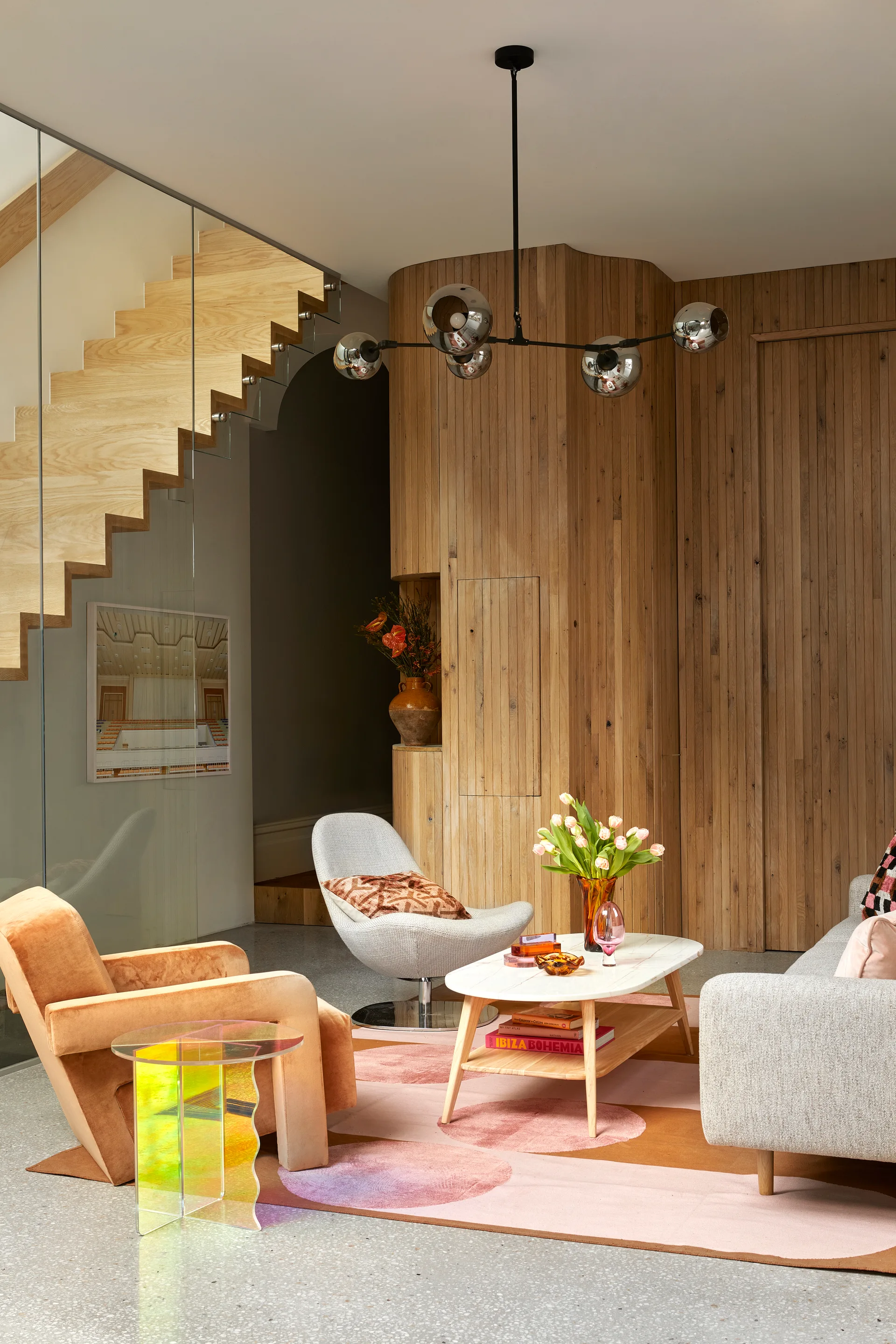 A contemporary living room featuring an orange armchair, a grey sofa, and a marble coffee table in front of a curved wood slat wall and floating staircase.