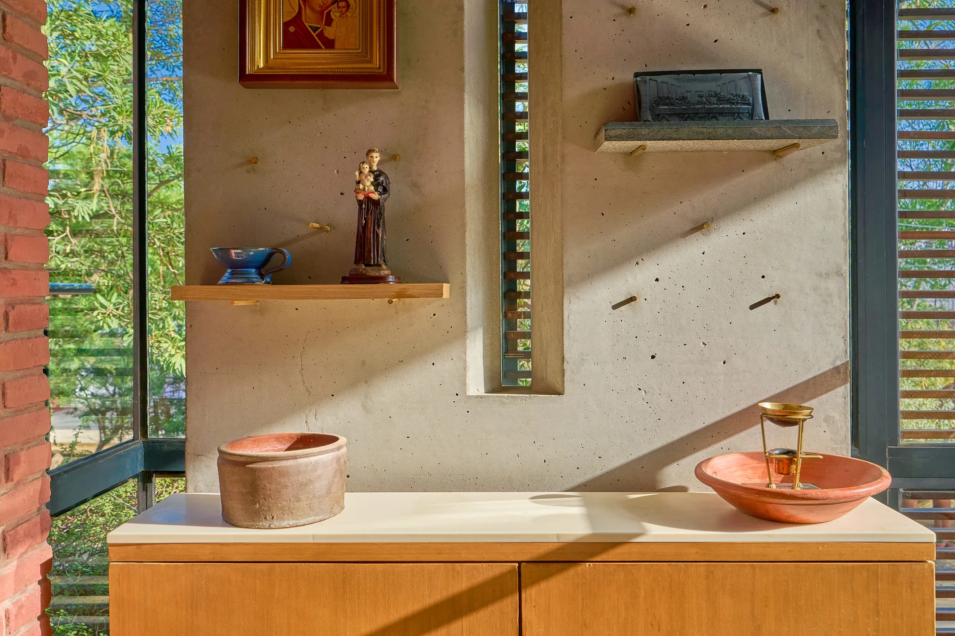 A smooth concrete wall with a narrow vertical window features two small floating shelves and a wooden console cabinet holding clay pottery.