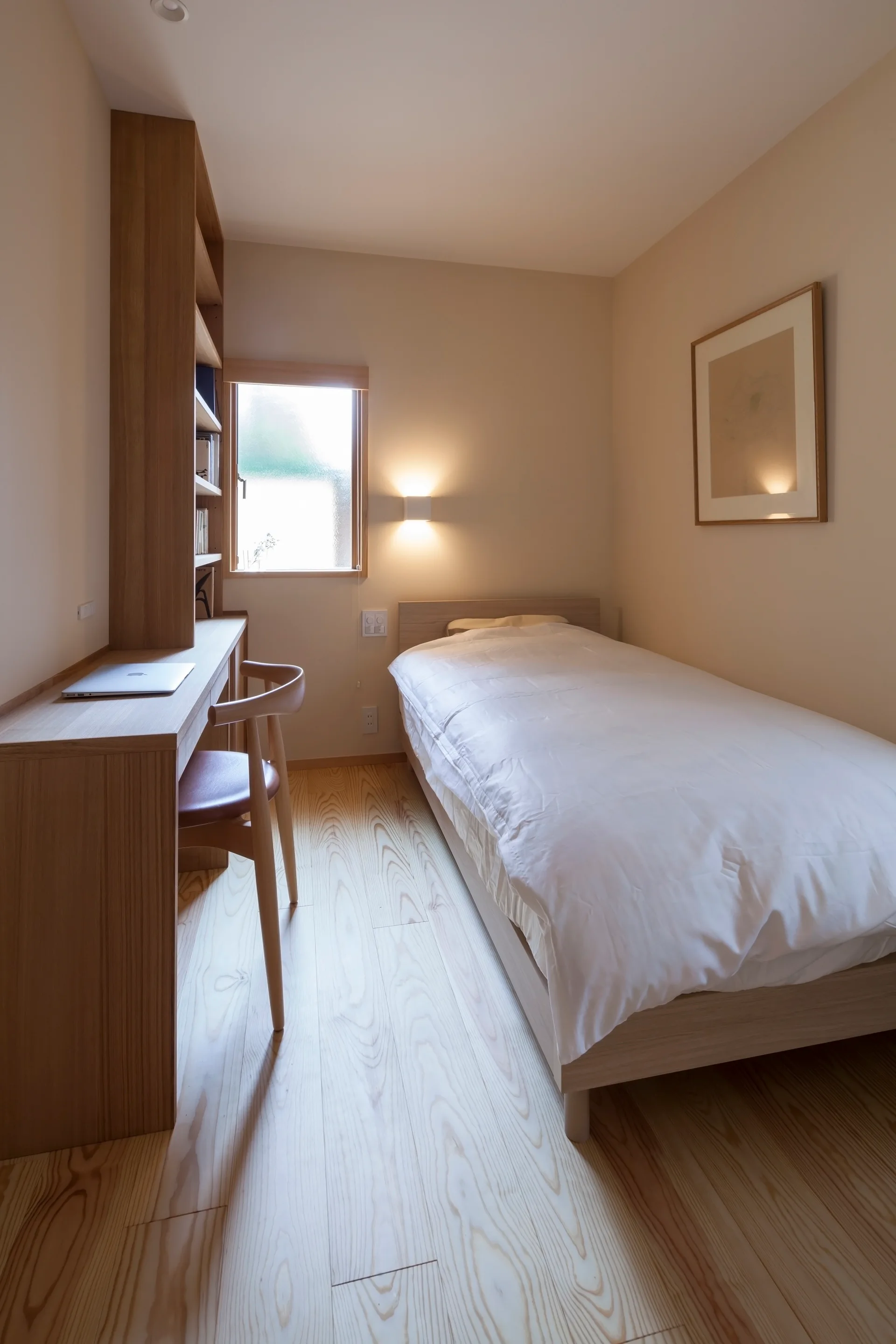 A small minimalist bedroom featuring a single bed with white bedding, a built-in wooden desk, and light wood flooring.