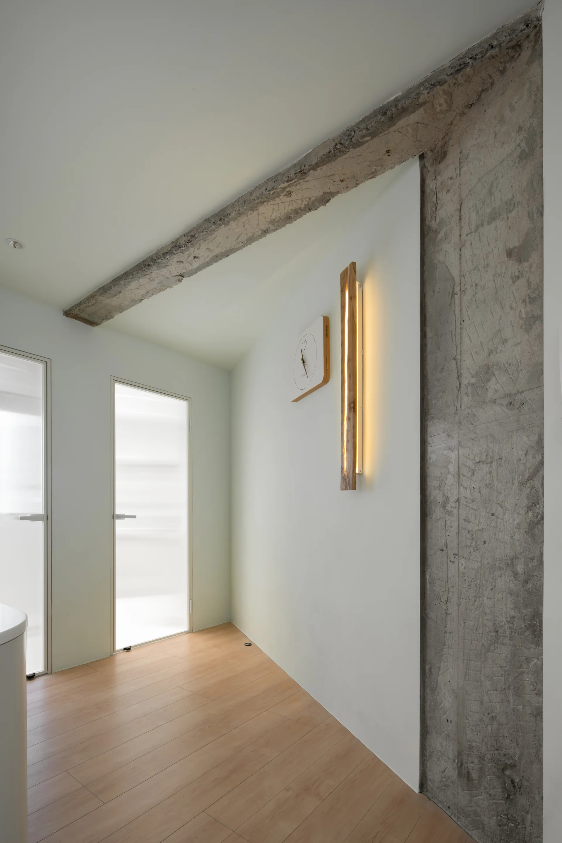 A minimalist hallway features light wood floors, an exposed concrete column and beam, frosted glass doors, and a glowing vertical wood sconce.
