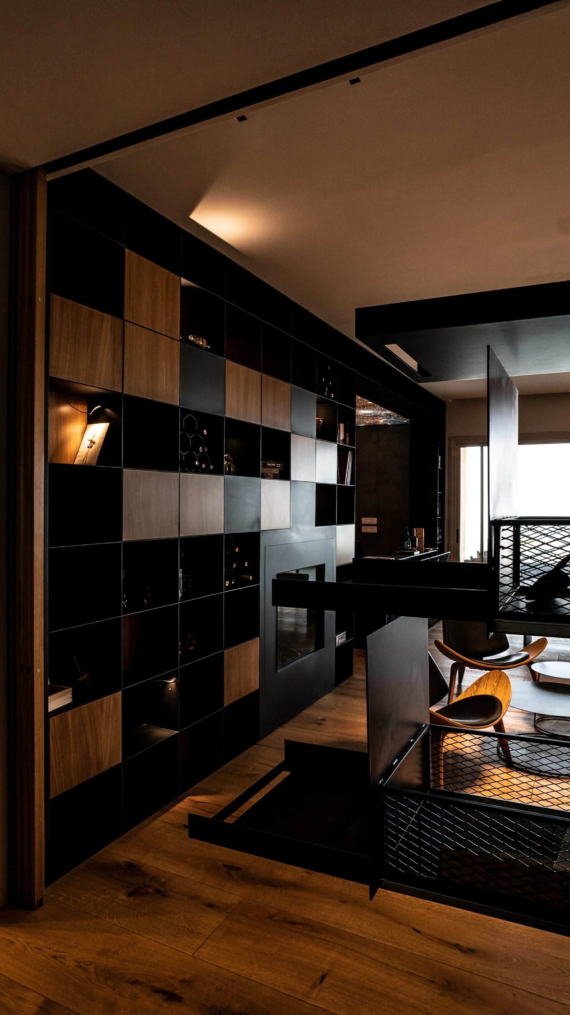 A modern living space features a custom floor-to-ceiling grid shelving unit with black and wood panels, an integrated fireplace, and wooden armchairs.