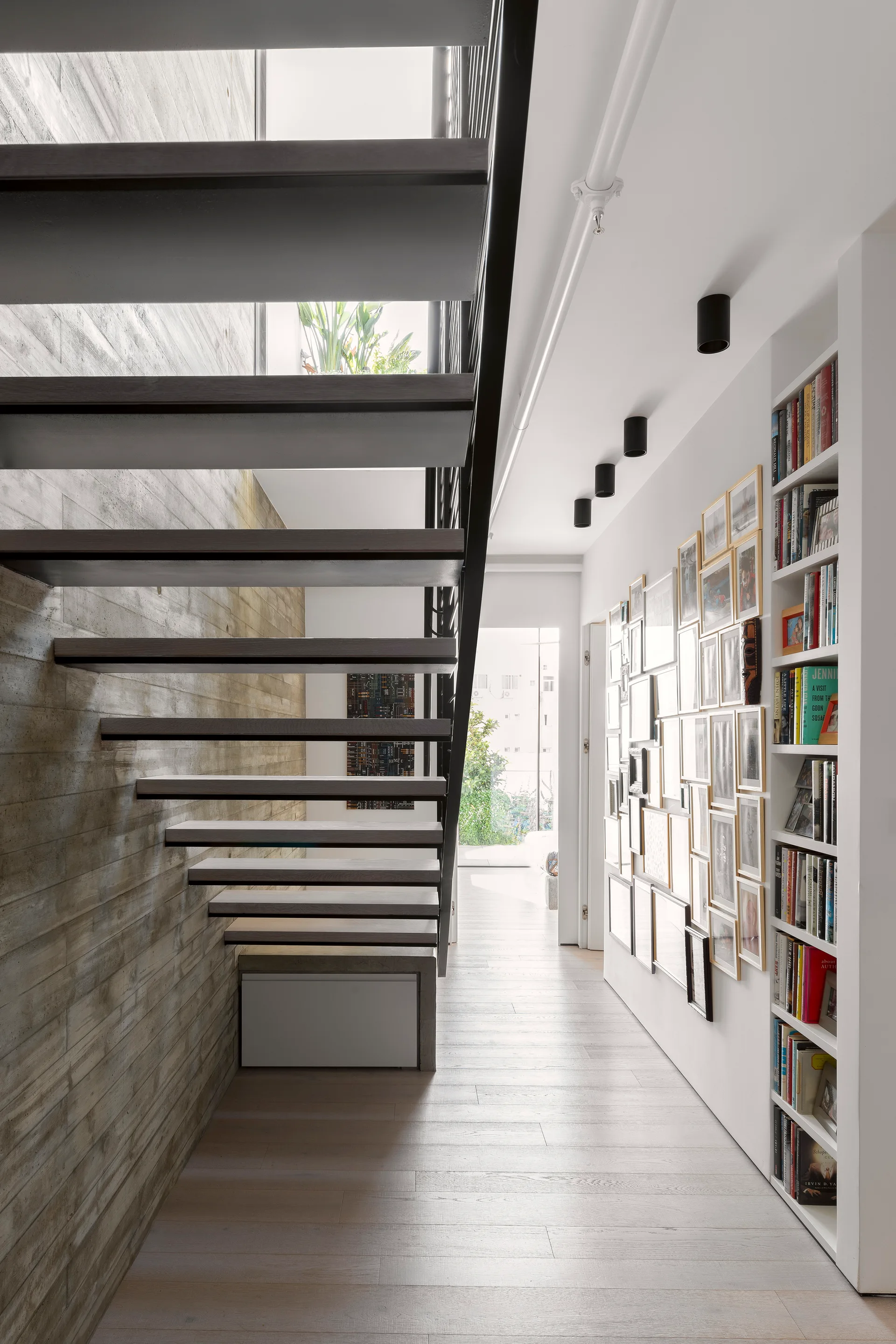 A floating wood staircase stands next to a textured concrete wall opposite a hallway with a gold-framed gallery wall and built-in bookshelf.