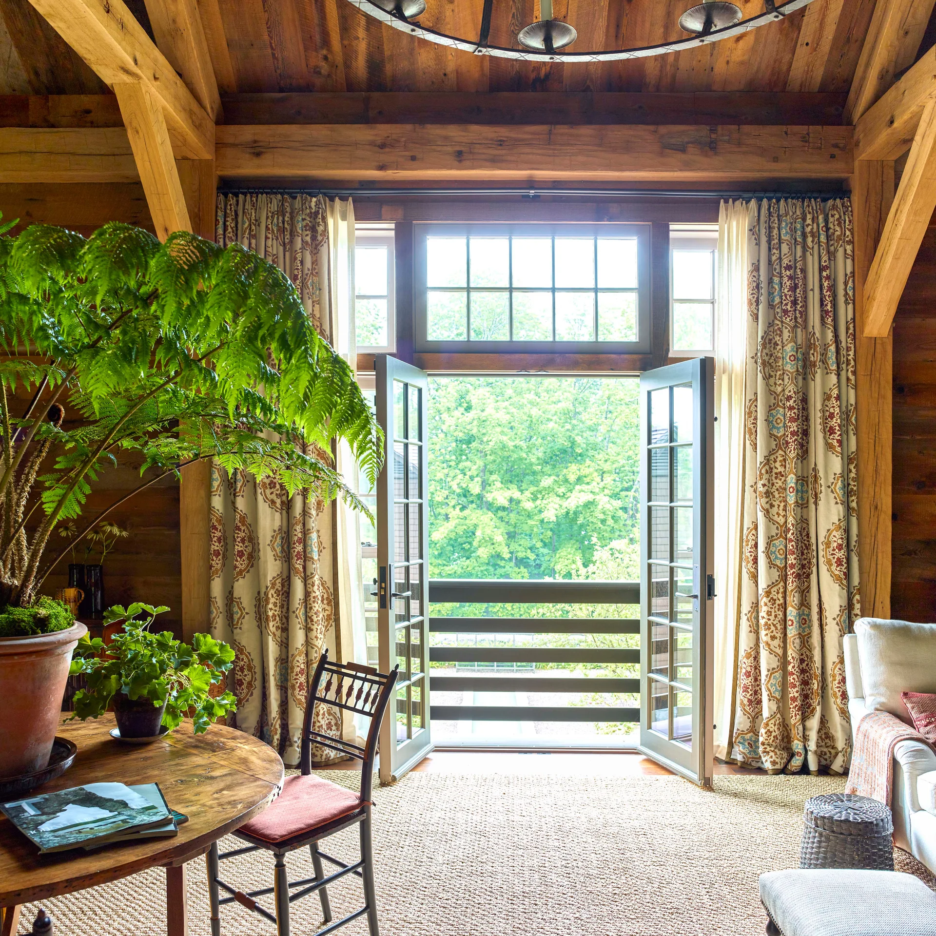 A rustic wooden living room features exposed timber beams, open French doors framing a lush green view, and patterned curtains.