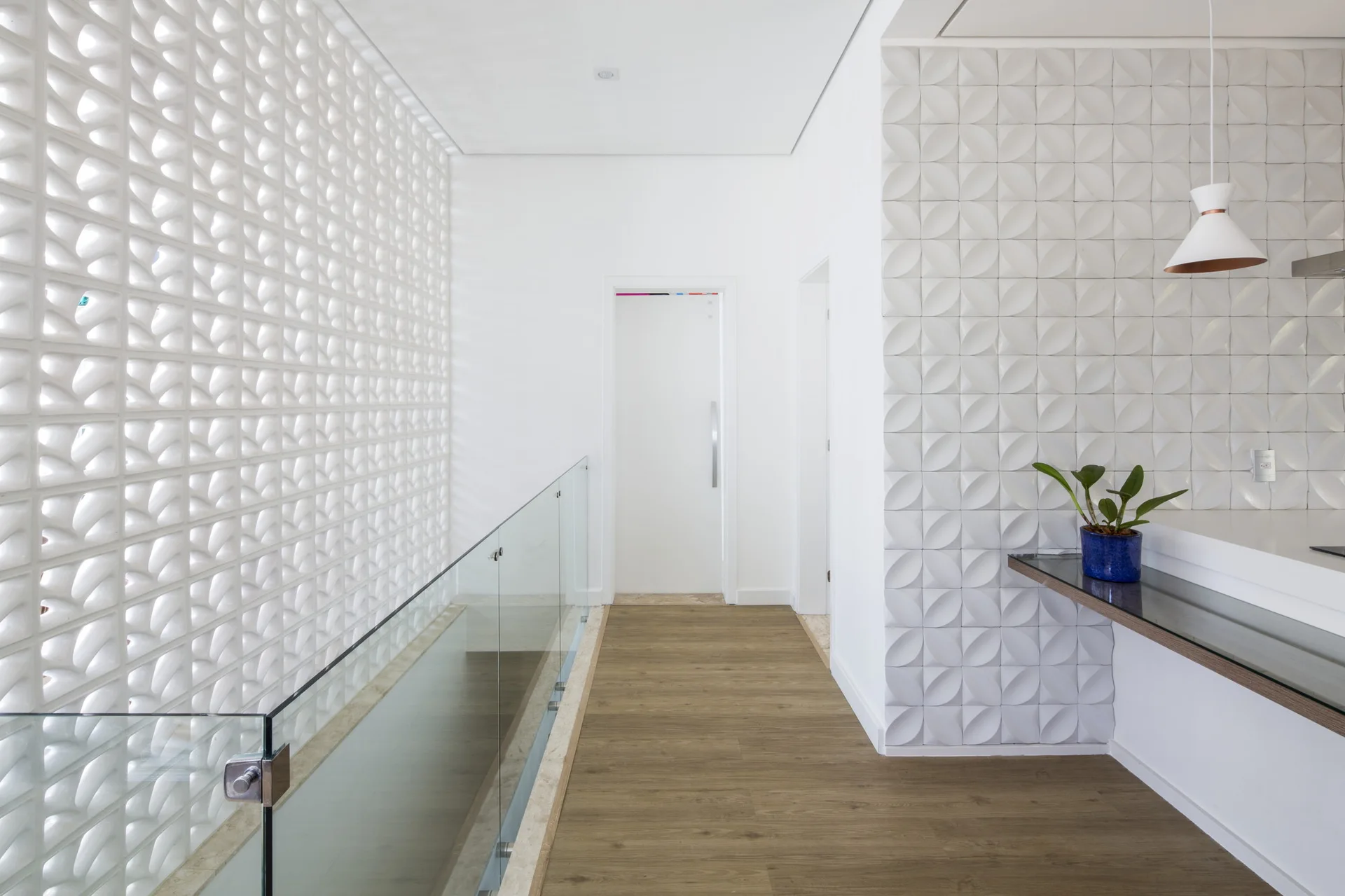 A bright hallway landing features a white breeze block wall, a glass railing, wooden flooring, and a geometric textured tile accent wall.