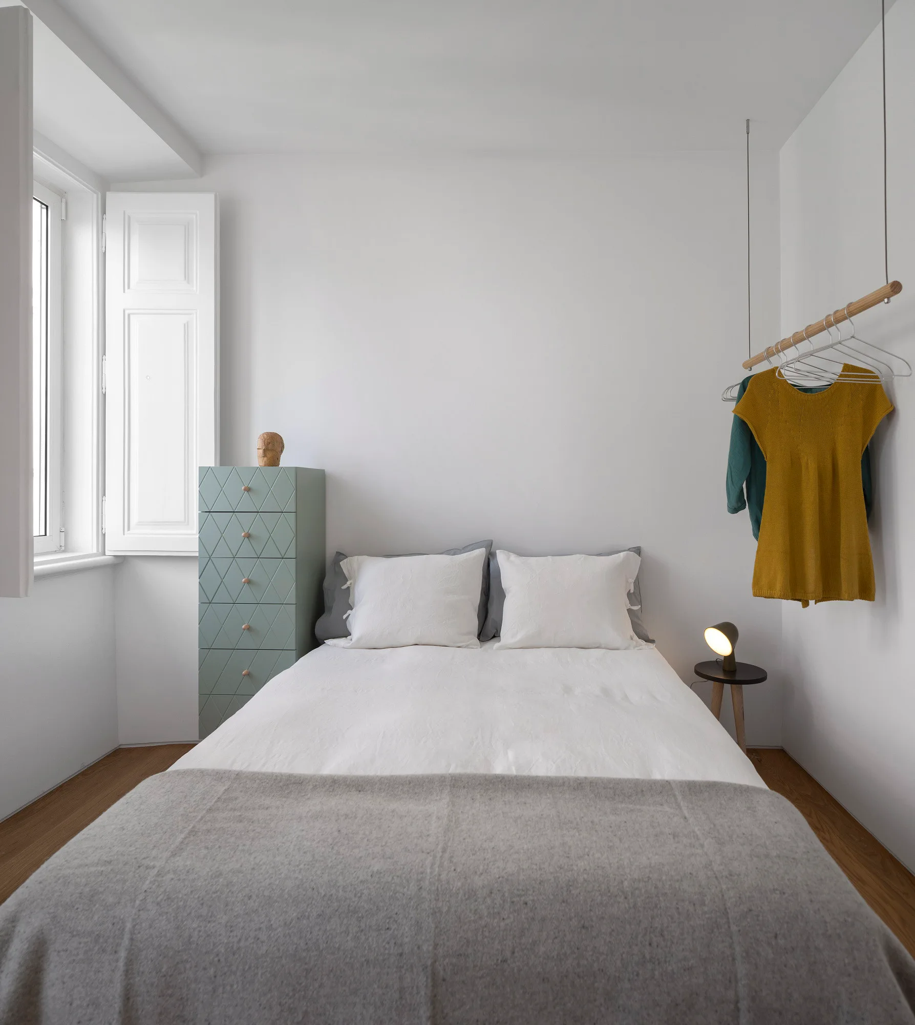 A minimalist bedroom featuring a bed with white bedding, a tall geometric sage green chest of drawers, and a ceiling-hung wooden clothing rod.