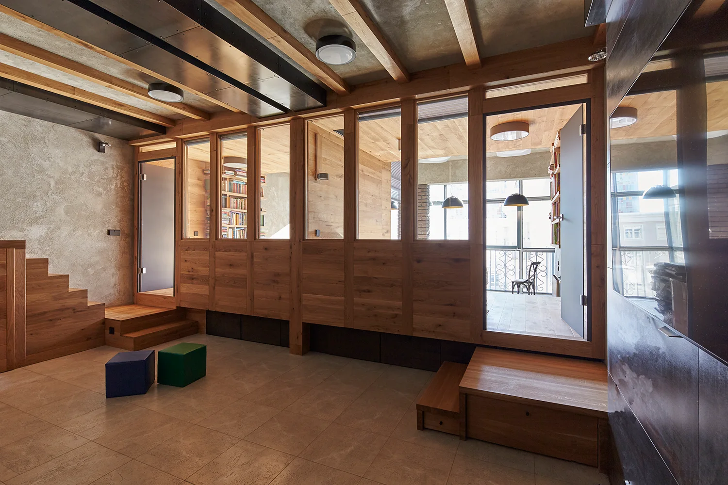 A spacious room features a large wood and glass partition wall, exposed ceiling beams over concrete, and a tiled floor with colorful cube seats.
