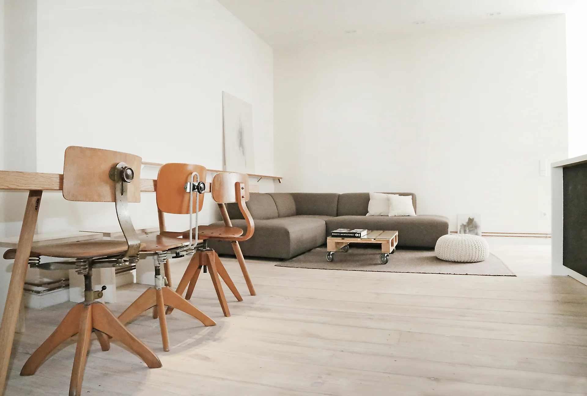 Three vintage wood swivel chairs stand on light wood flooring in a minimalist white living room with a grey sectional sofa and a pallet coffee table.