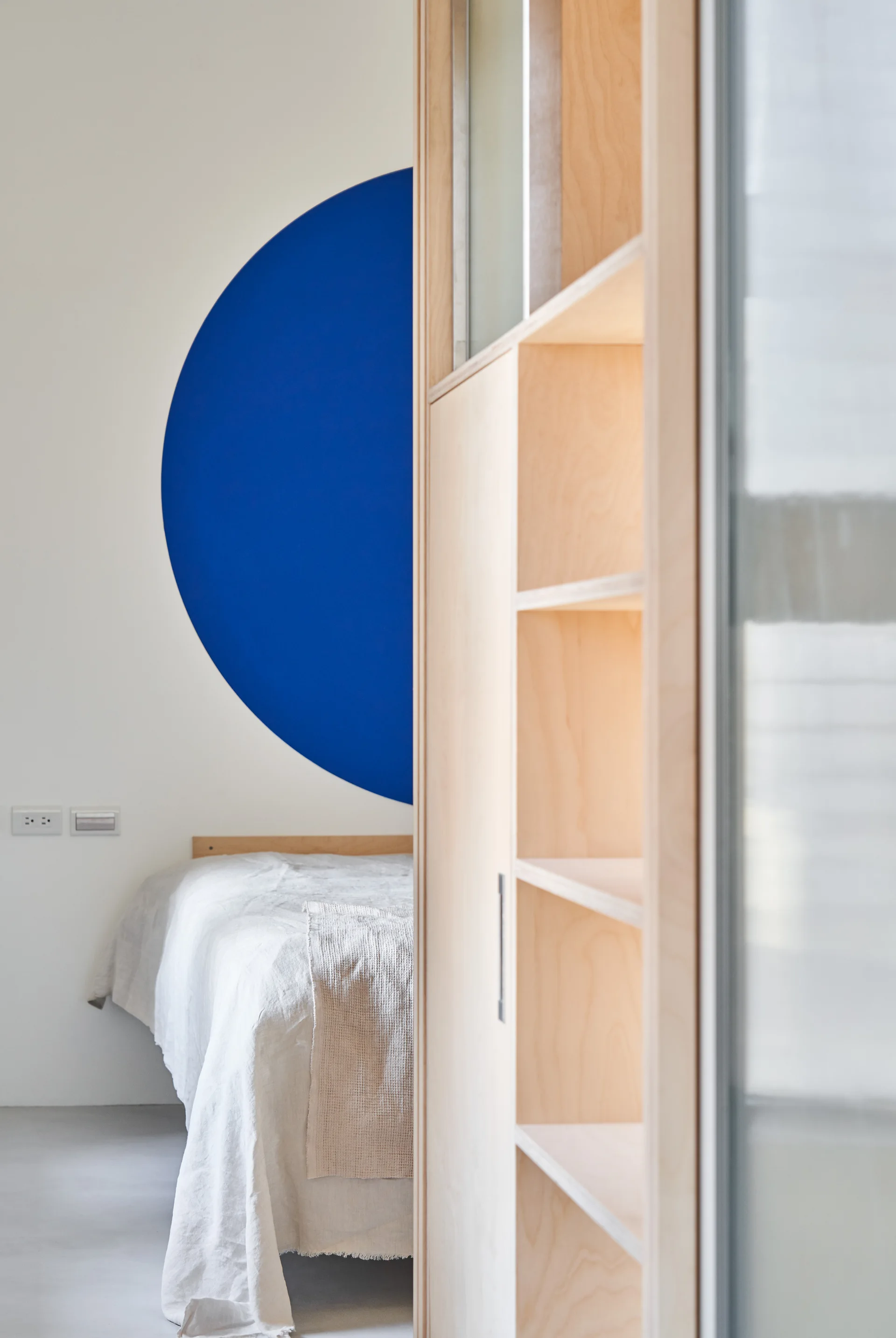 A minimalist bedroom features a bed with neutral linens next to a tall, light wood plywood cabinet against a white wall with a painted blue circle.