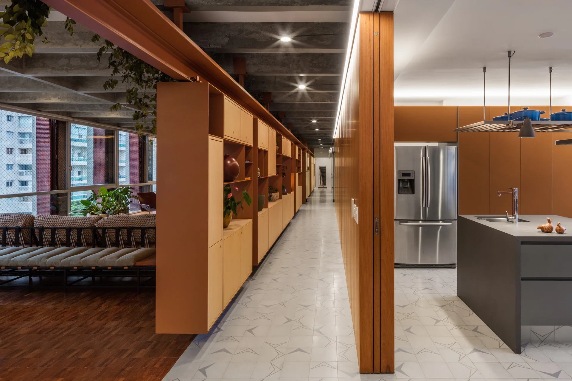 A modern open-concept kitchen and hallway featuring a long wood partition storage wall, grey kitchen island, and exposed concrete ceiling beams.