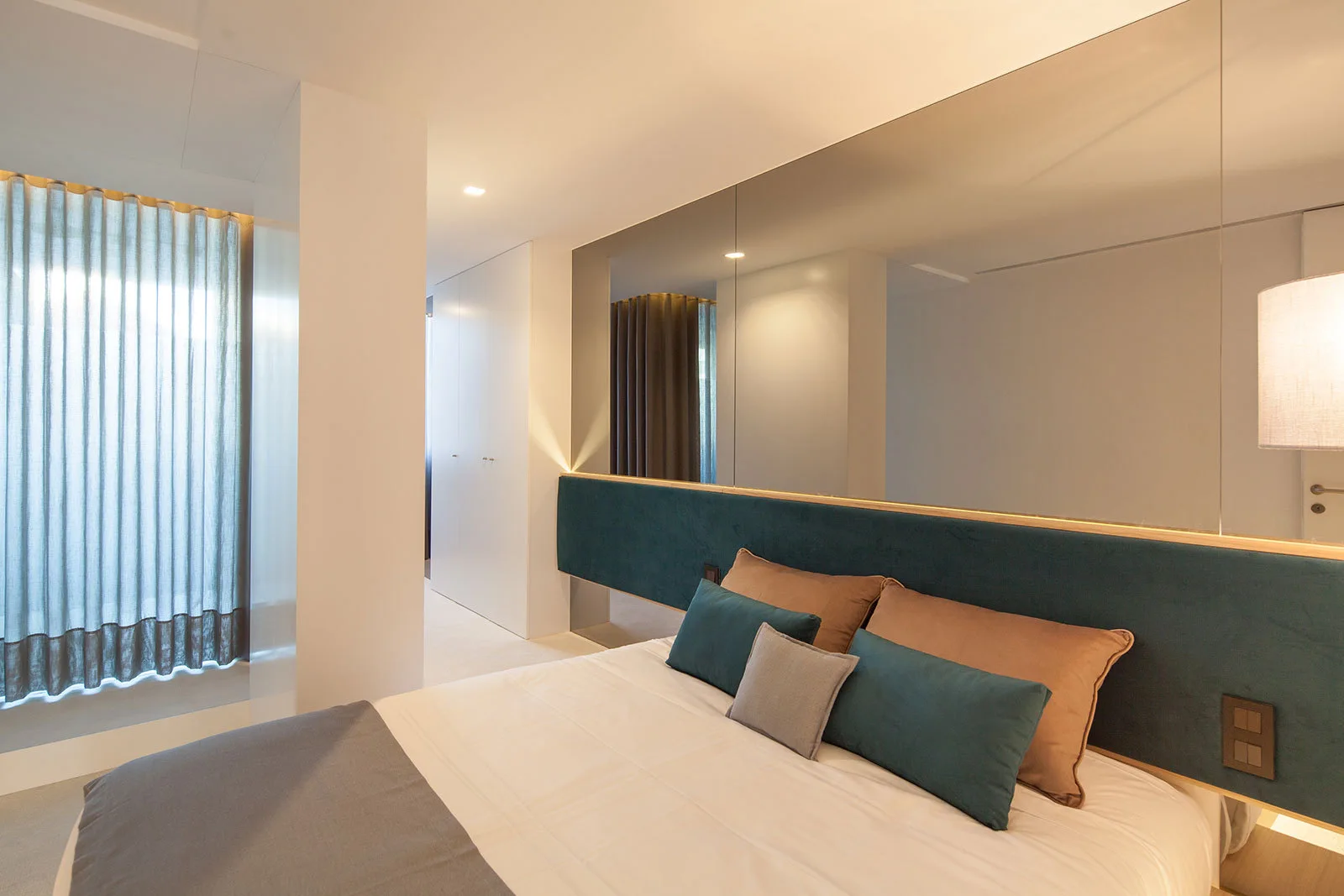 A modern bedroom features a large bed with teal and tan pillows, a low teal upholstered headboard, and a large mirrored wall panel above it.