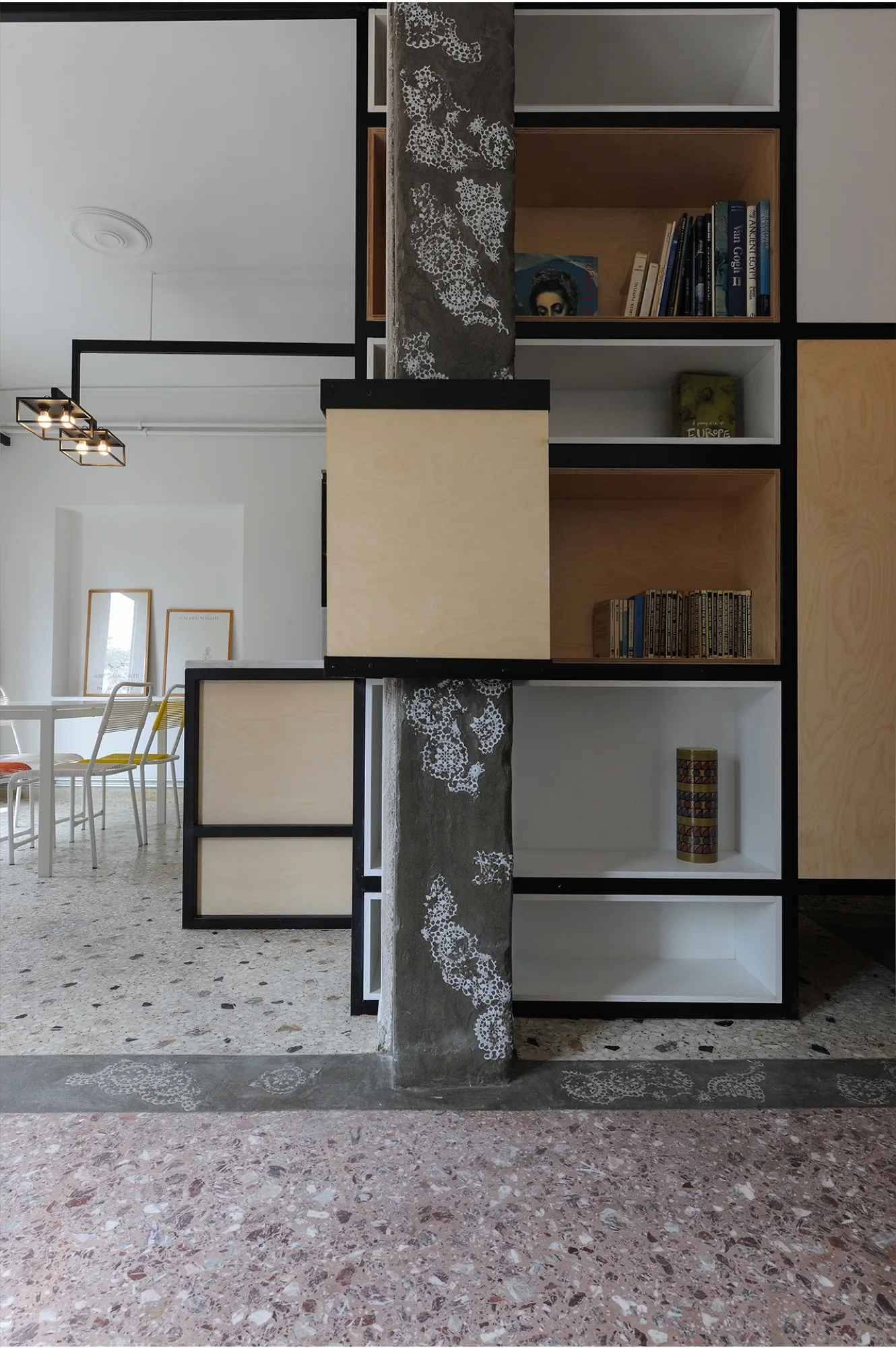 A modern room features a custom plywood shelving unit integrated with a concrete column, dining table, and terrazzo flooring.