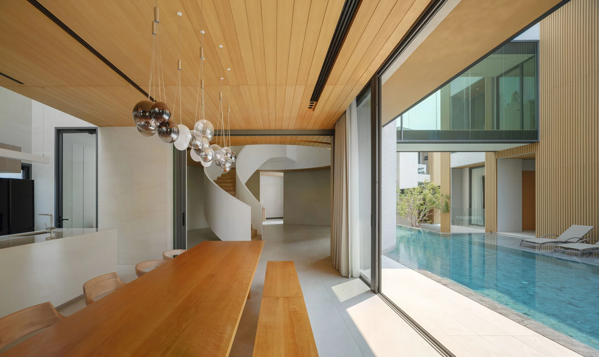 A modern dining room with a long wood table and bench features a wood-paneled ceiling and sliding glass doors opening to an outdoor pool.