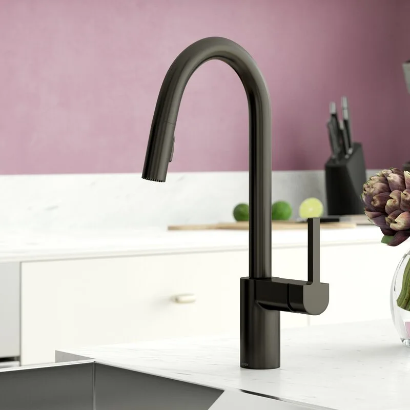 A matte black arched kitchen faucet stands on a white veined marble countertop against a dusty pink wall in a clean kitchen setting.
