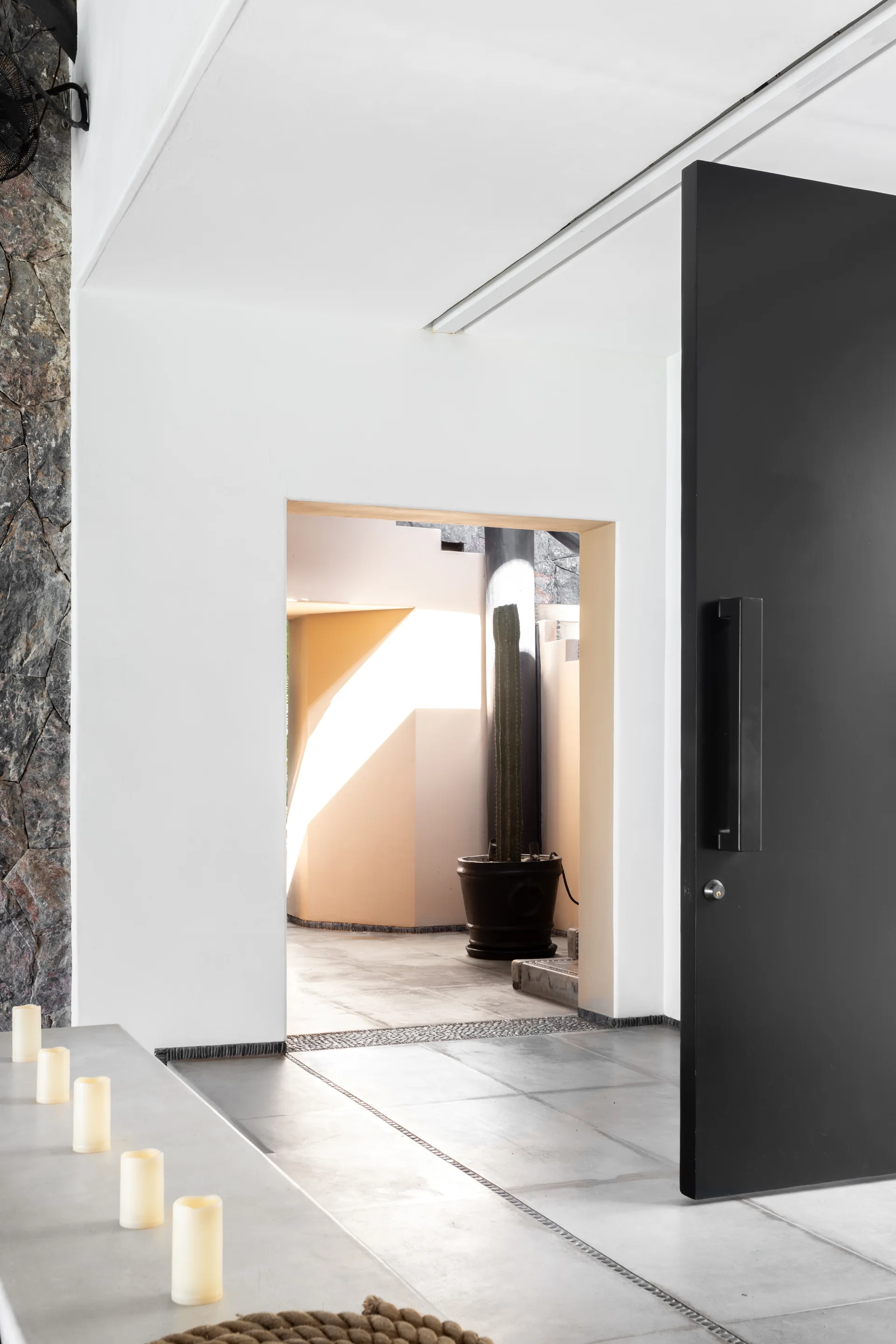 A minimalist entryway with a large black door, smooth grey tiled floors, and a framed opening leading to a sunlit courtyard with a cactus.
