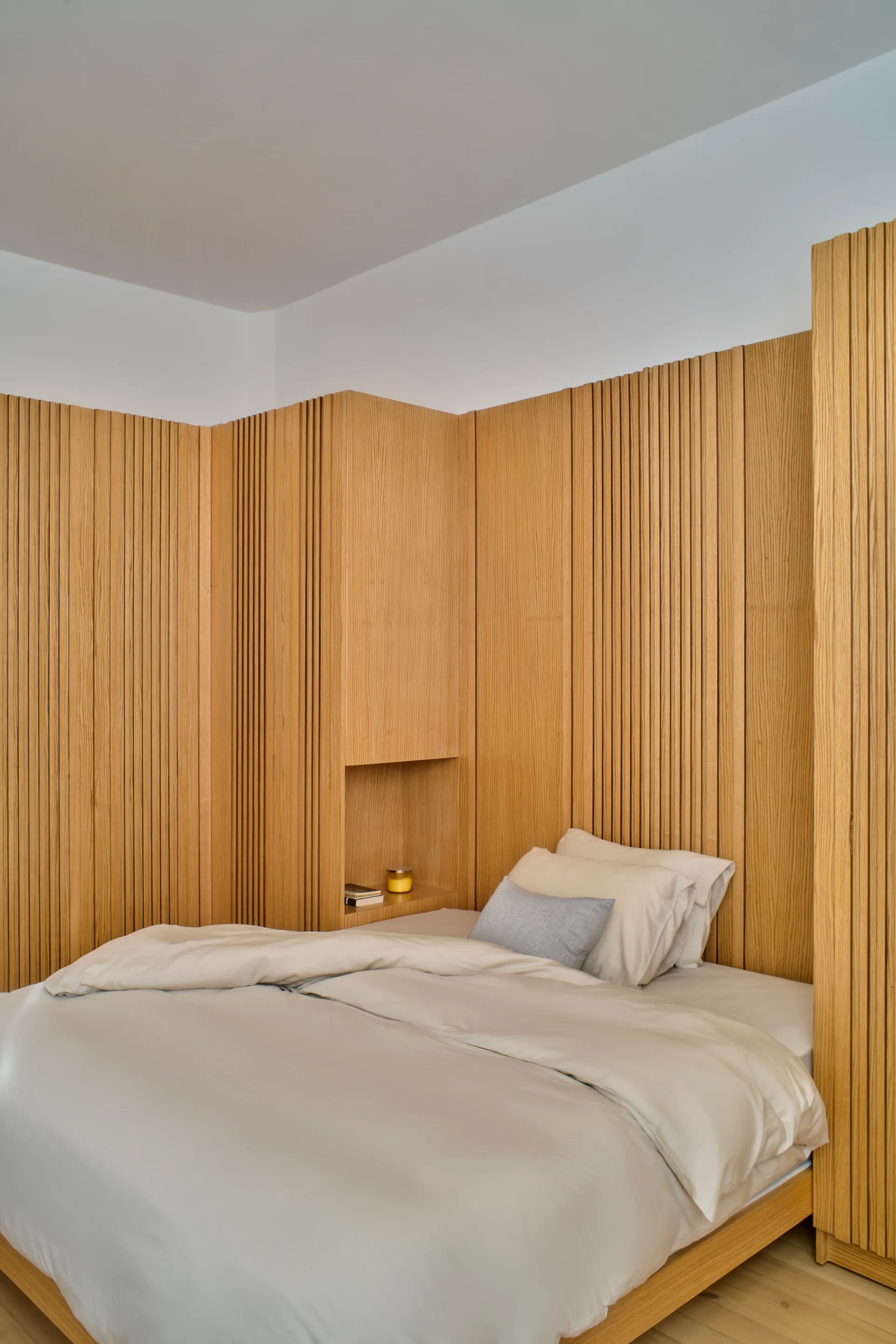 A low platform bed with neutral sheets rests against custom vertical wood slat paneling featuring a small built-in display niche.