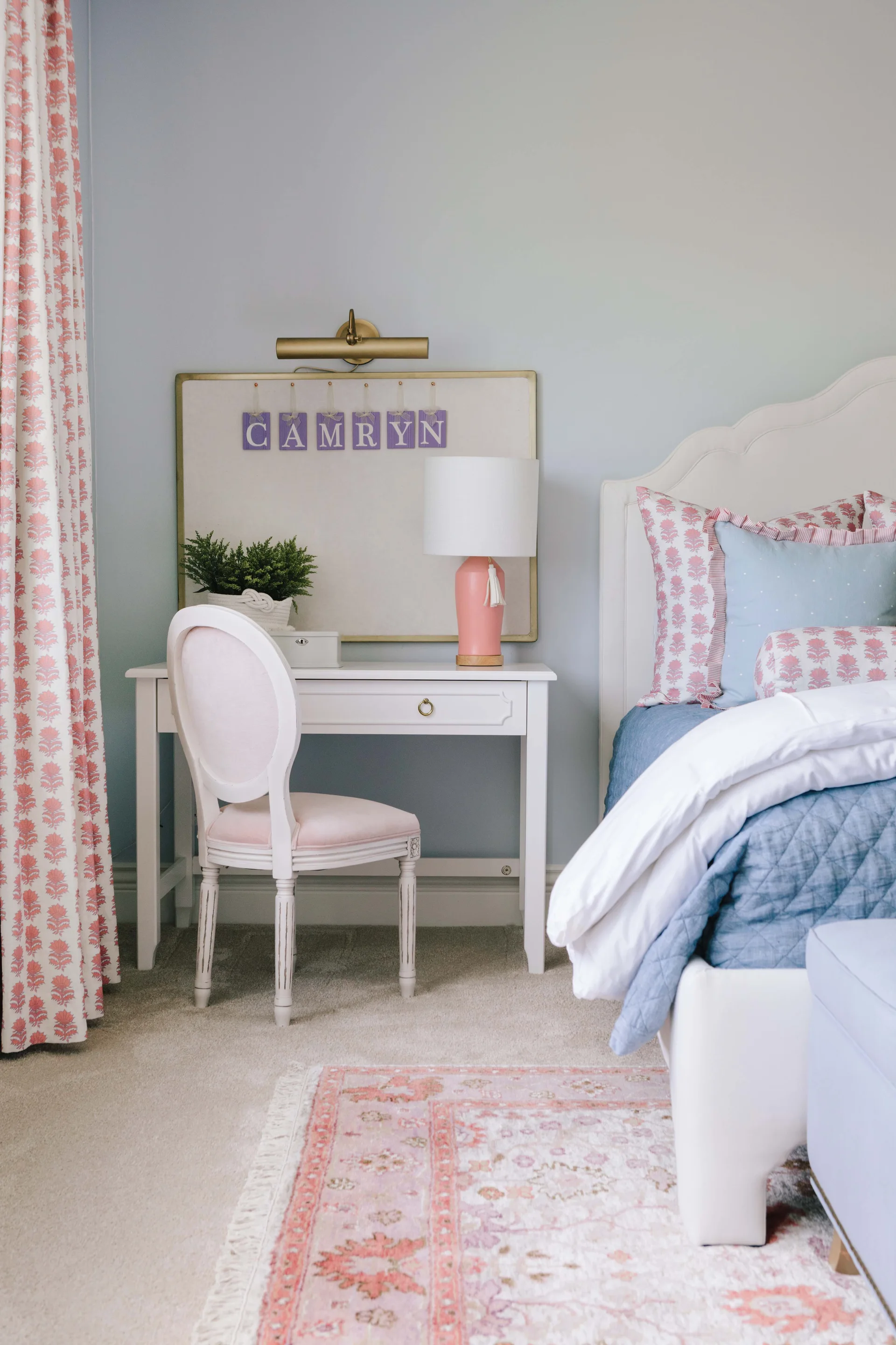 A kids bedroom featuring a white writing desk with a pink lamp, a medallion chair, and a bed with a scalloped headboard and blue quilt.