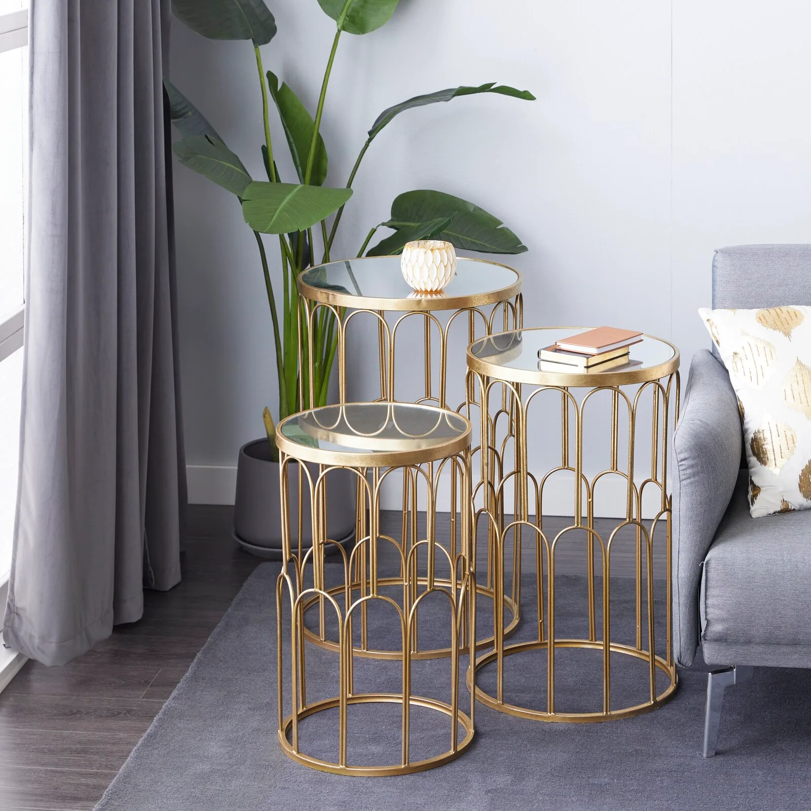 Three gold metal nesting tables with round glass tops stand on a grey area rug next to a grey fabric sofa and a large potted plant.