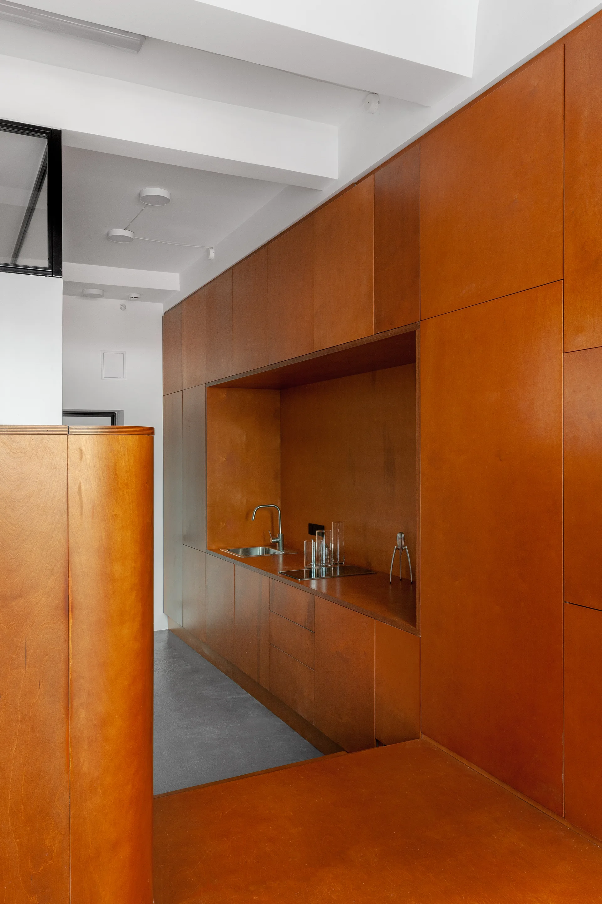 A minimalist kitchen features floor to ceiling warm wood cabinetry, a recessed sink countertop niche, gray concrete floors, and exposed white ceiling beams.