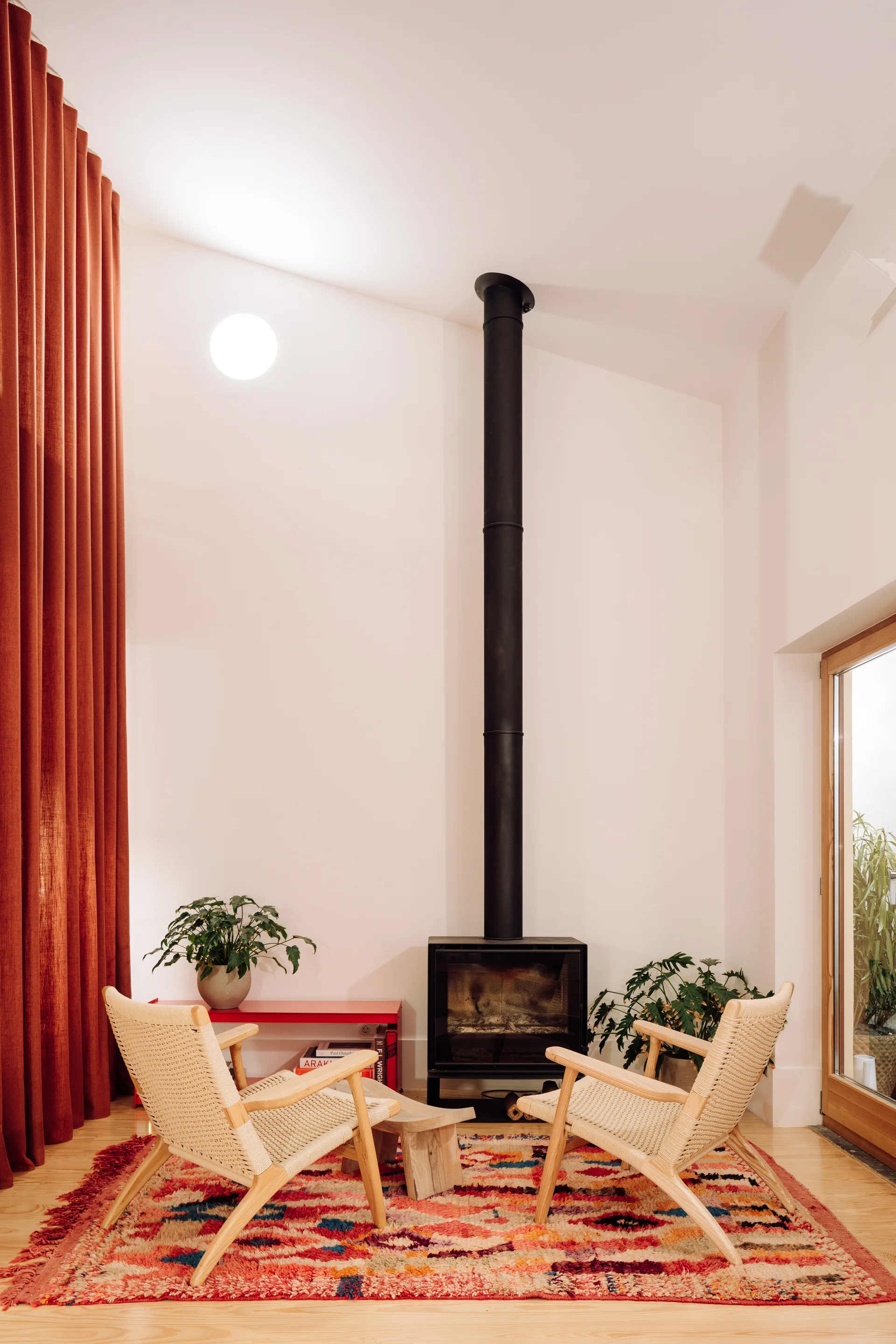 Two light wood armchairs with woven seats face a tall, black freestanding fireplace on a colorful patterned rug in a room with white walls.