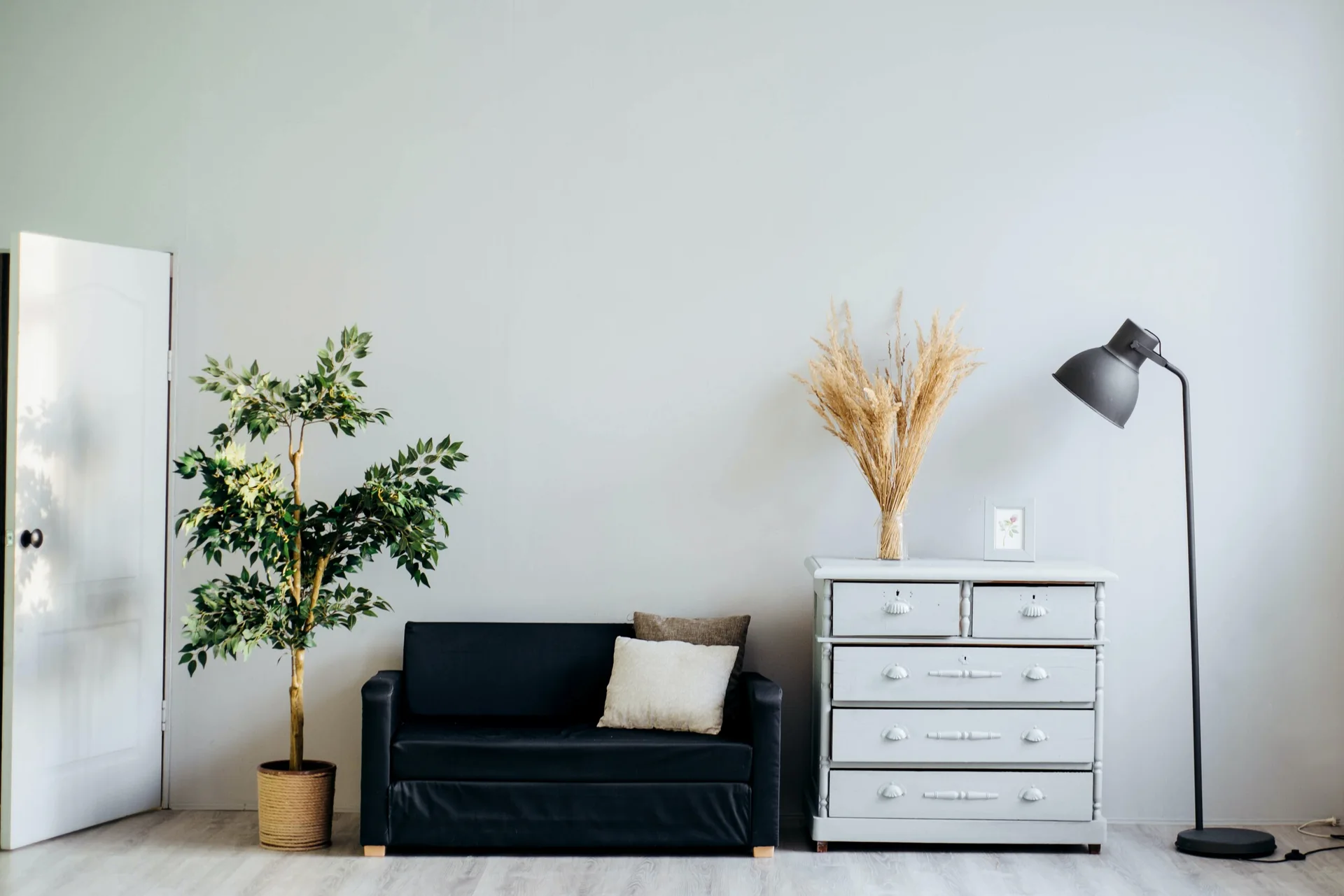 A small black sofa sits next to a light grey painted dresser with dried grass, flanked by a potted green tree and a black floor lamp against a pale grey wall.