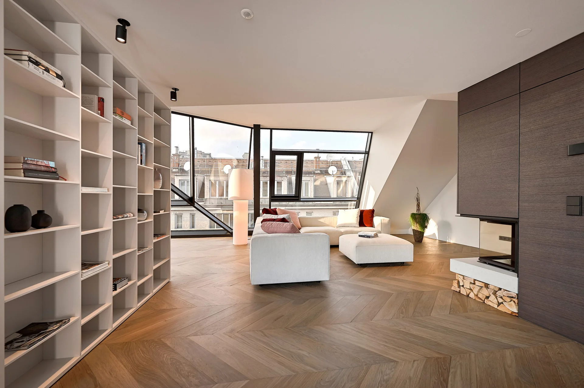 A modern attic living room featuring a white modular sofa, custom bookshelves, chevron wood flooring, and a large angular window.