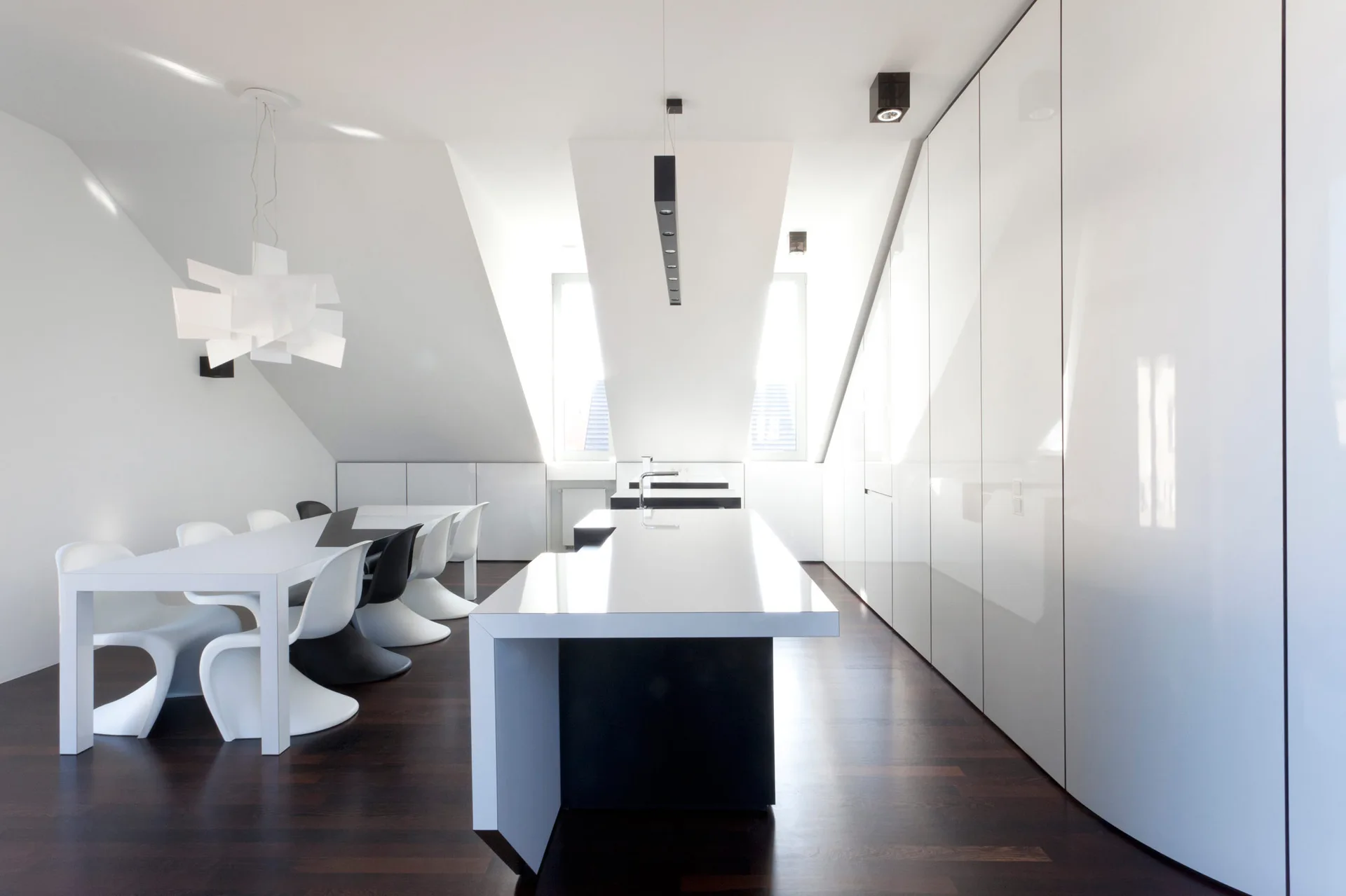 A minimalist kitchen under a sloped ceiling features a glossy white island, a dining table with sculptural chairs, and tall handleless cabinets.