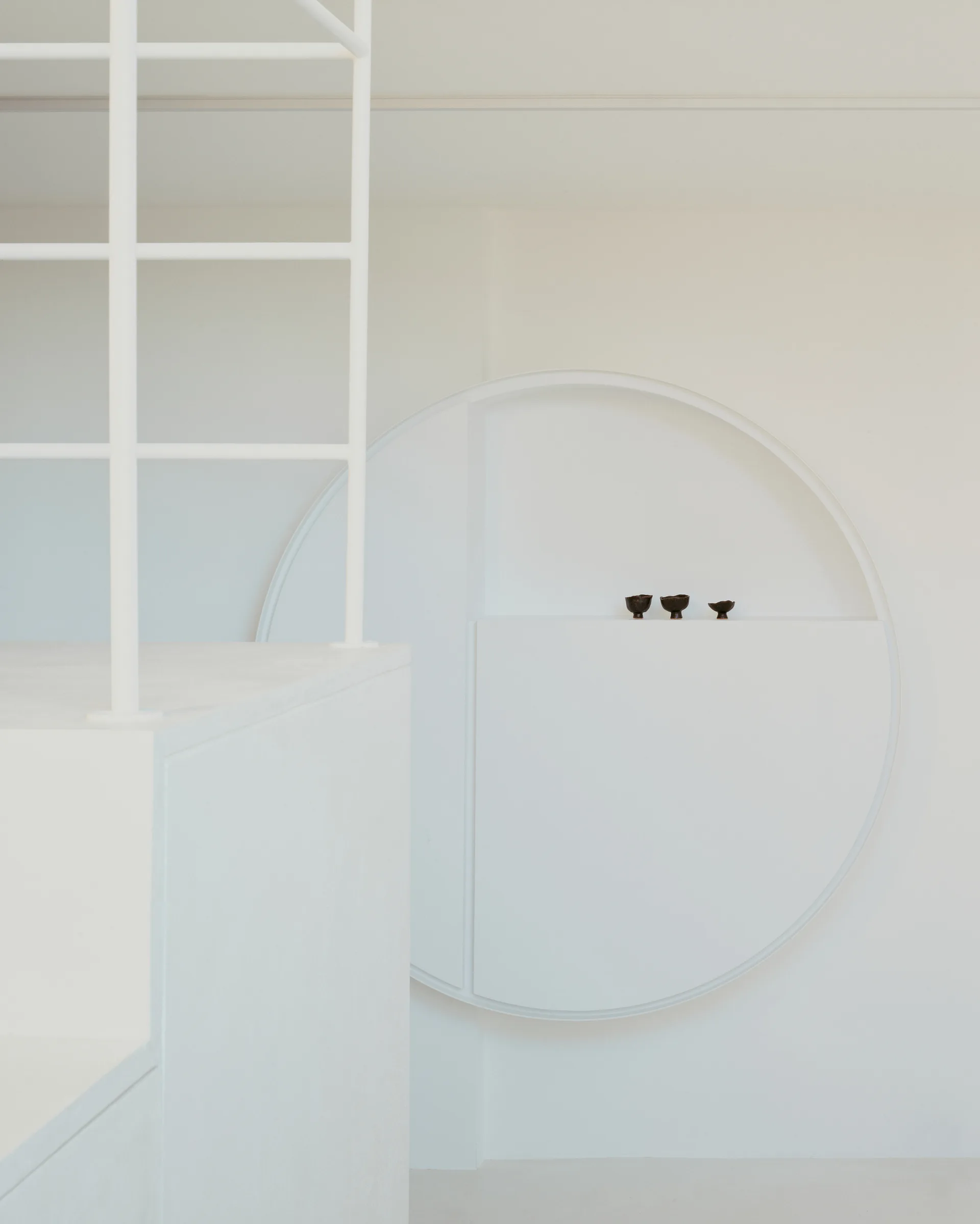 A minimalist white staircase with a grid railing stands next to a large recessed circular wall niche holding three tiny dark bowls.