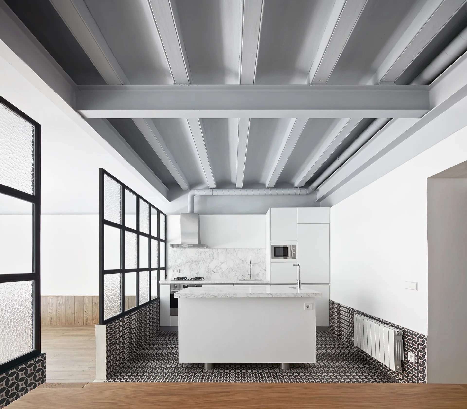 A modern white kitchen features a marble-topped island, patterned floor tiles, gray painted ceiling beams, and a black metal glass partition.