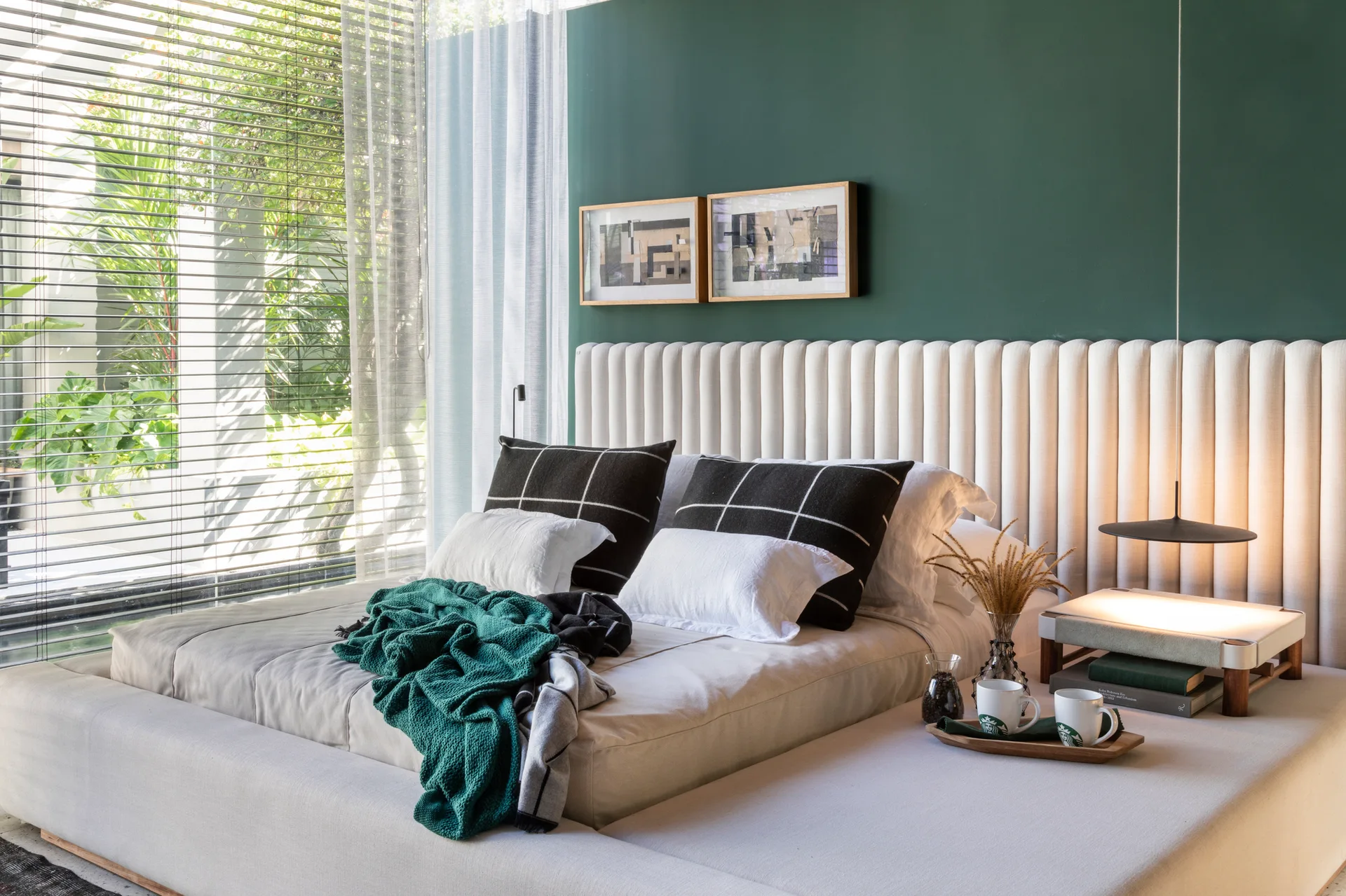 A modern bedroom features a low platform bed with a cream channel-tufted headboard against a forest green accent wall next to a large window.