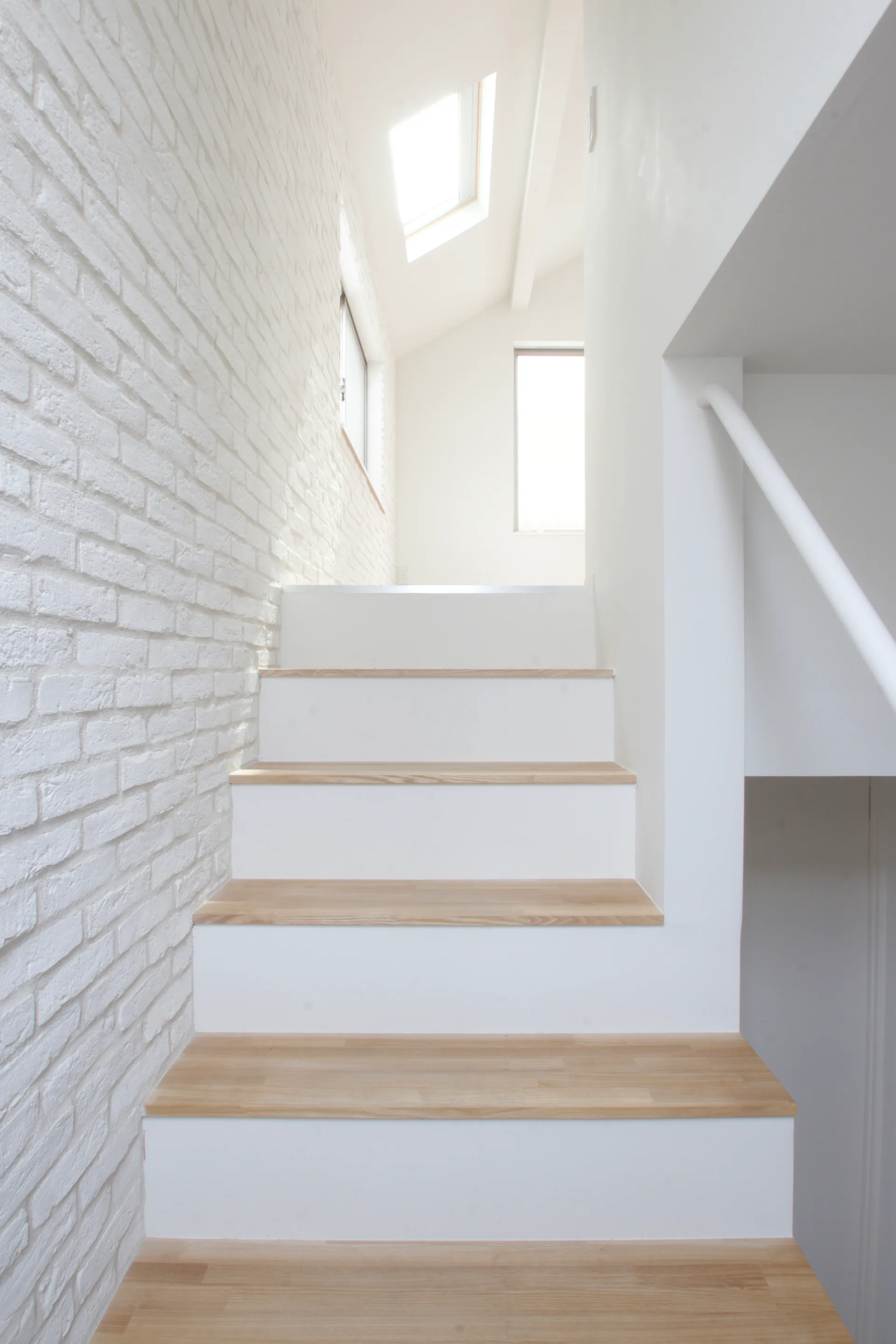 A minimalist staircase with light wood treads and white risers features a white painted brick accent wall under a bright ceiling skylight.