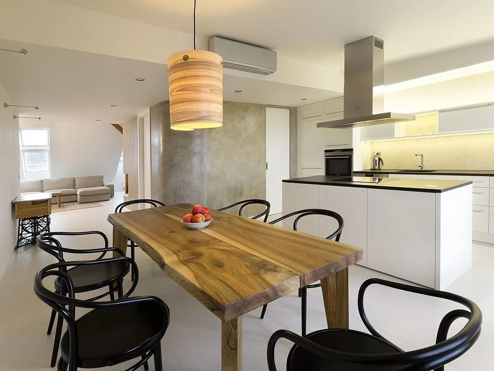 A modern open-plan kitchen and dining area featuring a live-edge wood table, black chairs, a white kitchen island, and a textured concrete wall.