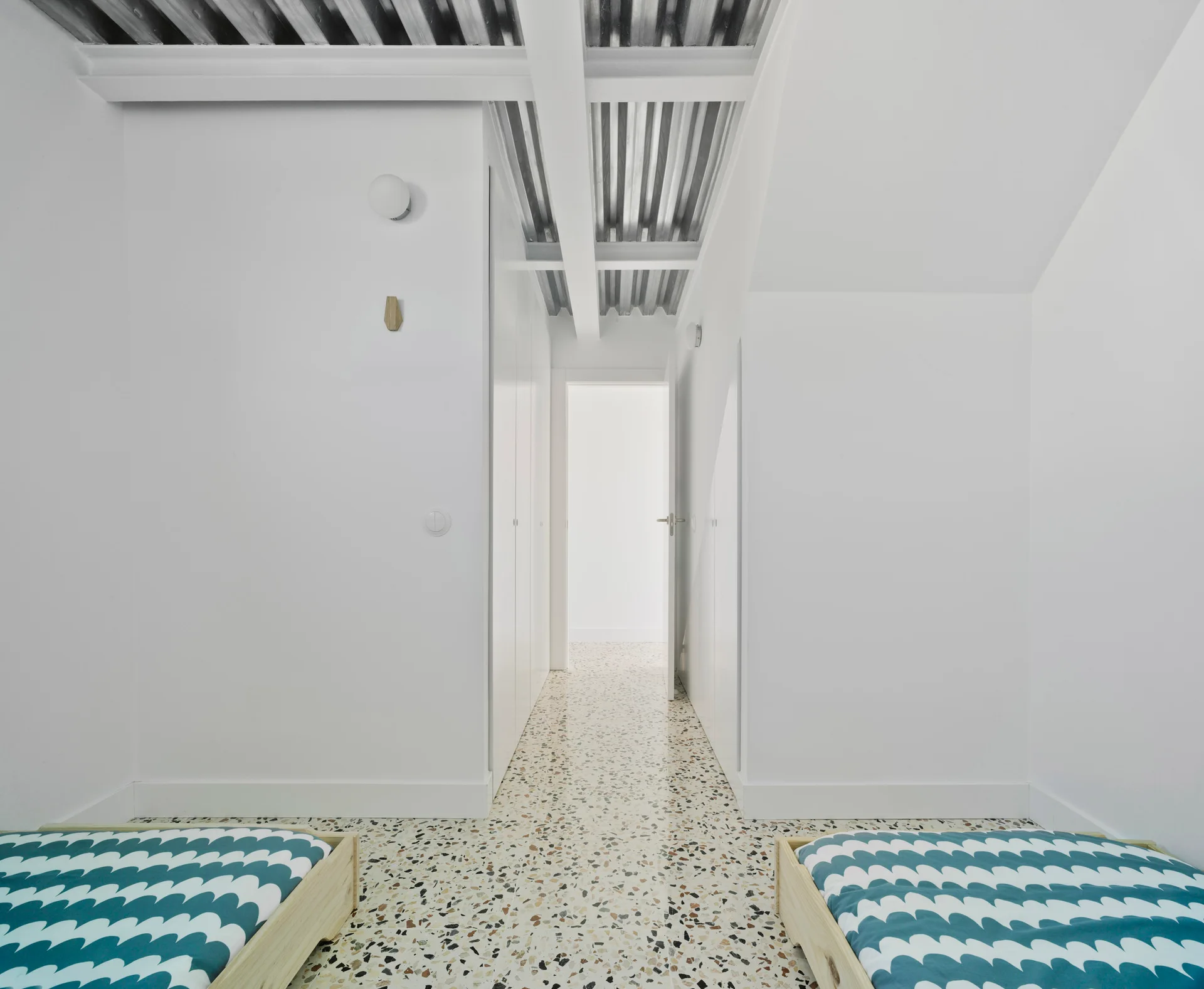 A white bedroom features twin beds, colorful terrazzo flooring, and an exposed corrugated metal ceiling with structural beams.