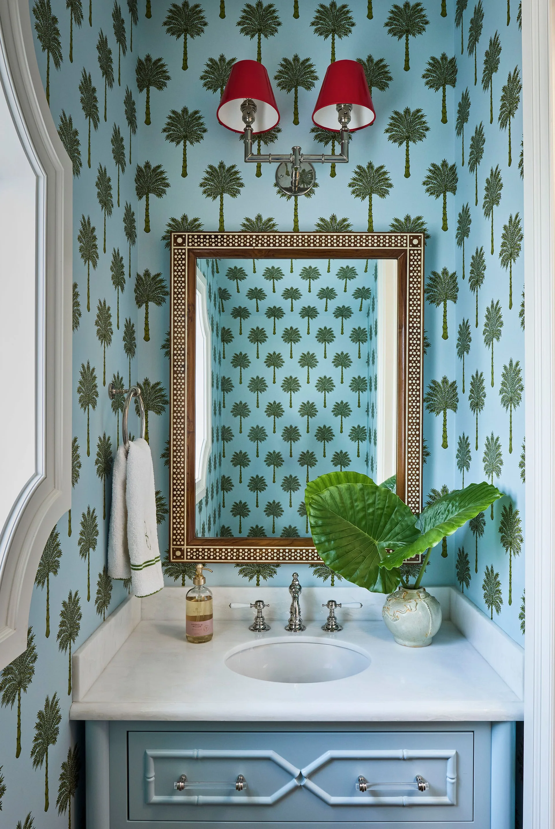 A small powder room with blue palm tree wallpaper, a custom blue vanity with a stone countertop, a patterned mirror, and a sconce with red shades.