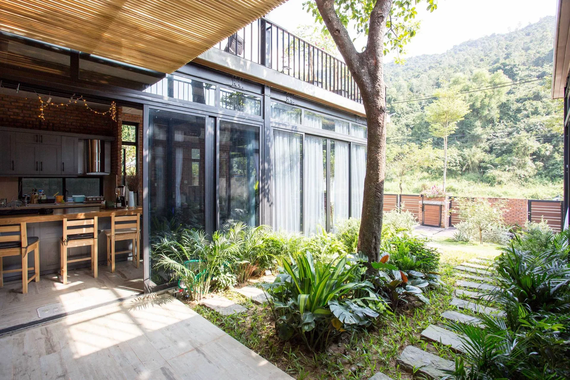 A kitchen with wooden island seating opens through large sliding glass doors onto a garden patio with stone stepping stones and lush green plants.