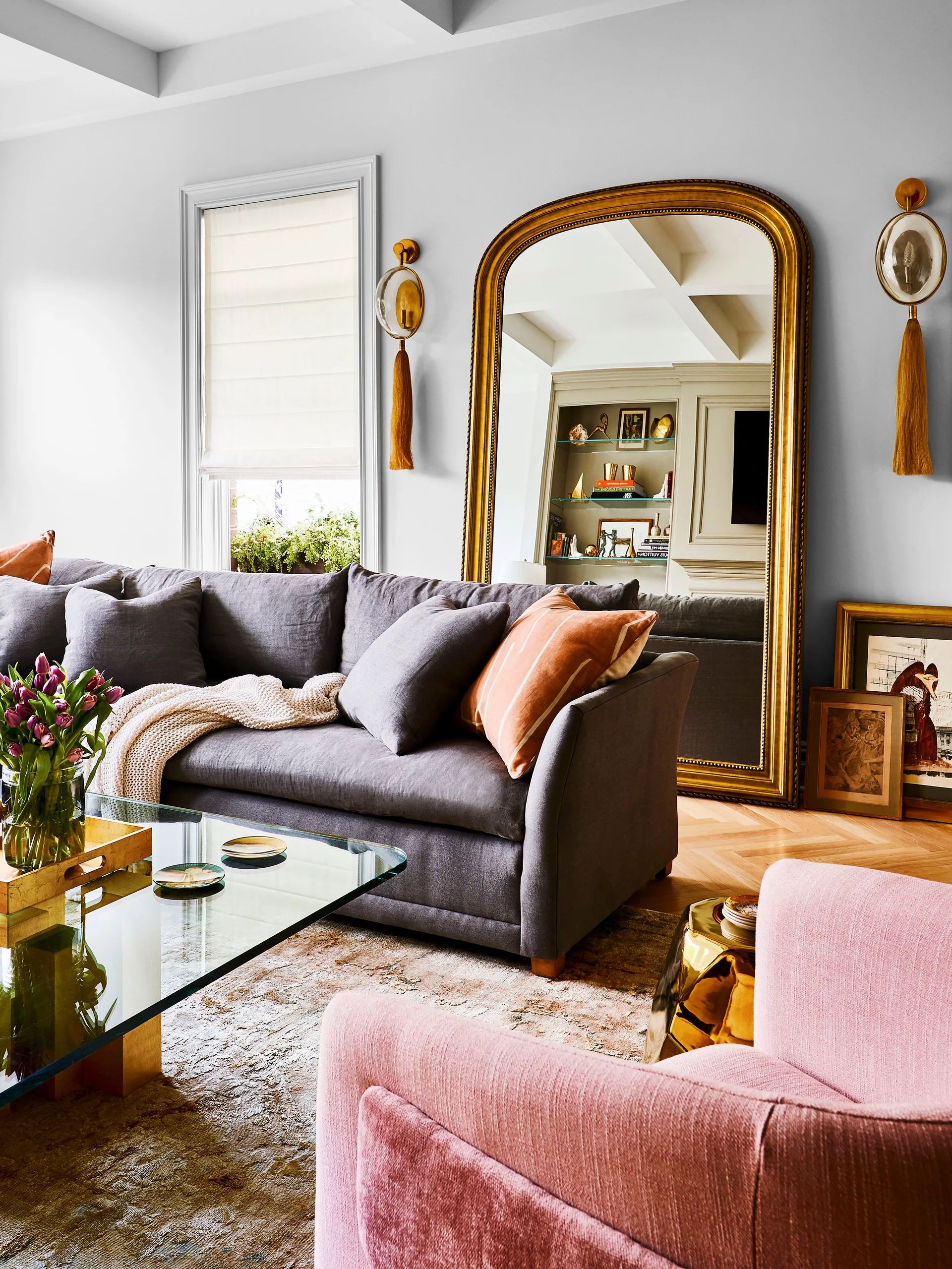 A living room featuring a grey sofa, a pink armchair, a glass coffee table, and a large gold-framed arched mirror leaning against the wall.