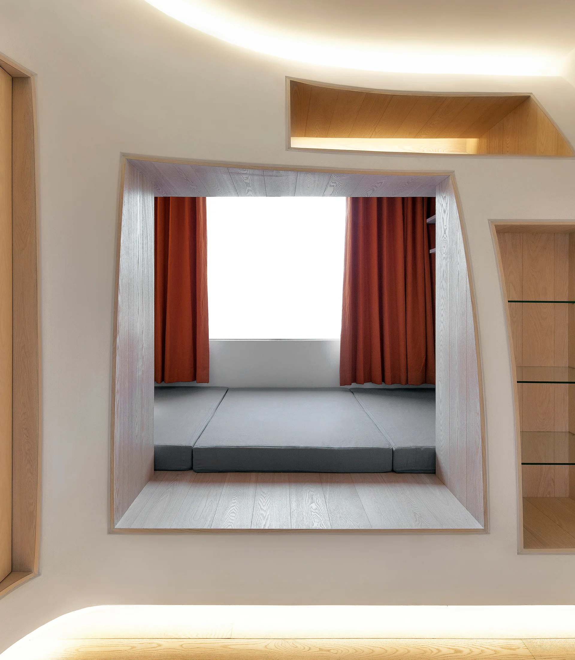 A minimalist recessed sleeping nook set into a curved white wall features a grey floor mattress, terracotta curtains, and integrated light wood shelving.
